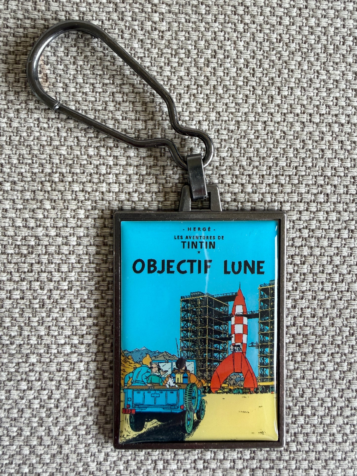 A rectangular keychain displaying the French book cover art for Hergé's Tintin adventure "Objectif Lune." The illustration shows Tintin, Captain Haddock, Snowy, and Professor Calculus watching from an open-top blue vehicle as the iconic red-and-white checkered moon rocket towers over a bustling launchpad against a backdrop of rugged mountains.
