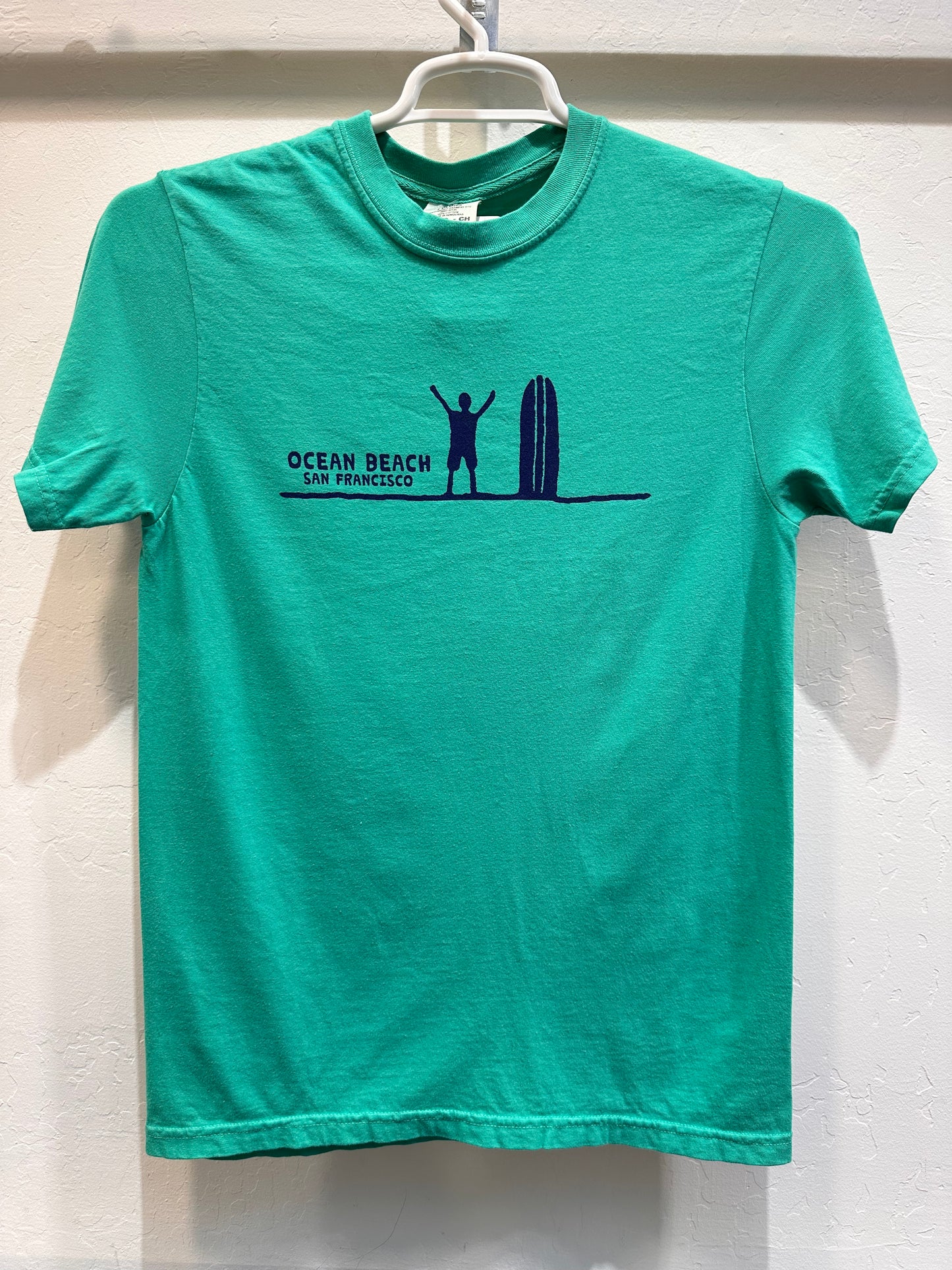 Front of a Kelly Green t-shirt displaying a minimalist navy blue graphic across the chest. It features a silhouette of a surfer standing with arms raised next to a vertical surfboard on a single horizon line, captioned with the words "OCEAN BEACH SAN FRANCISCO."