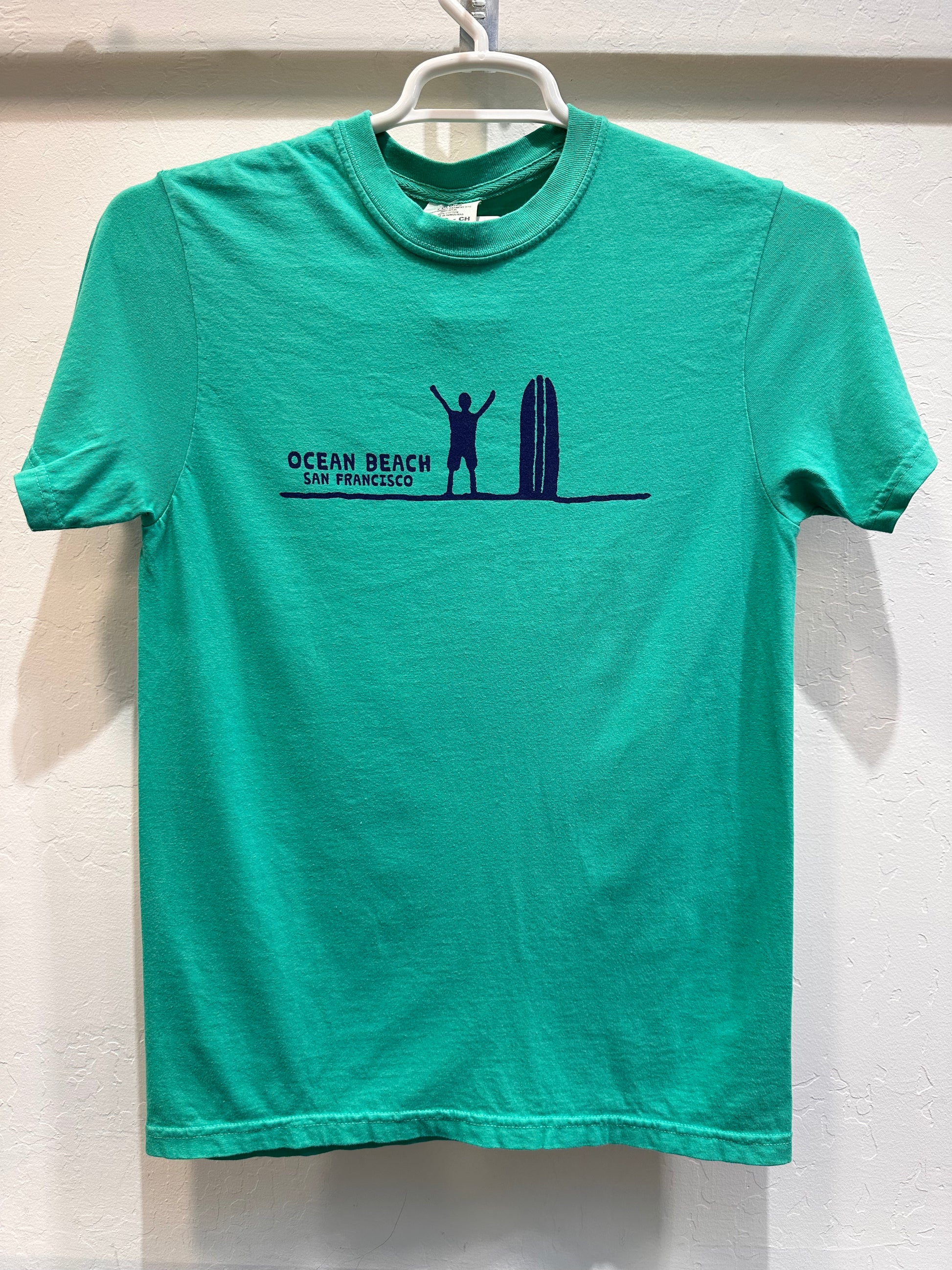 Front of a Kelly Green t-shirt displaying a minimalist navy blue graphic across the chest. It features a silhouette of a surfer standing with arms raised next to a vertical surfboard on a single horizon line, captioned with the words "OCEAN BEACH SAN FRANCISCO."