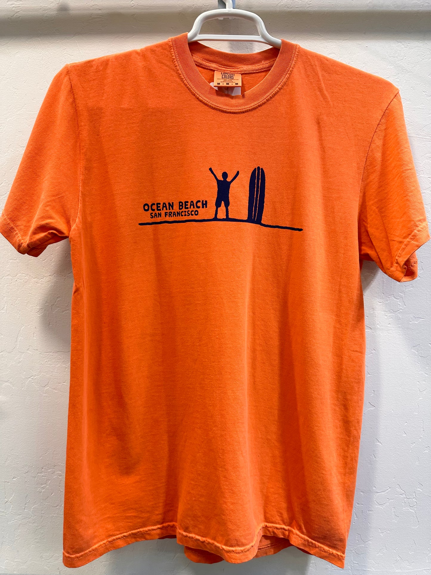 Front of a terracotta orange t-shirt displaying a minimalist navy blue graphic across the chest. It features a silhouette of a surfer standing with arms raised next to a vertical surfboard on a single horizon line, captioned with the words "OCEAN BEACH SAN FRANCISCO."