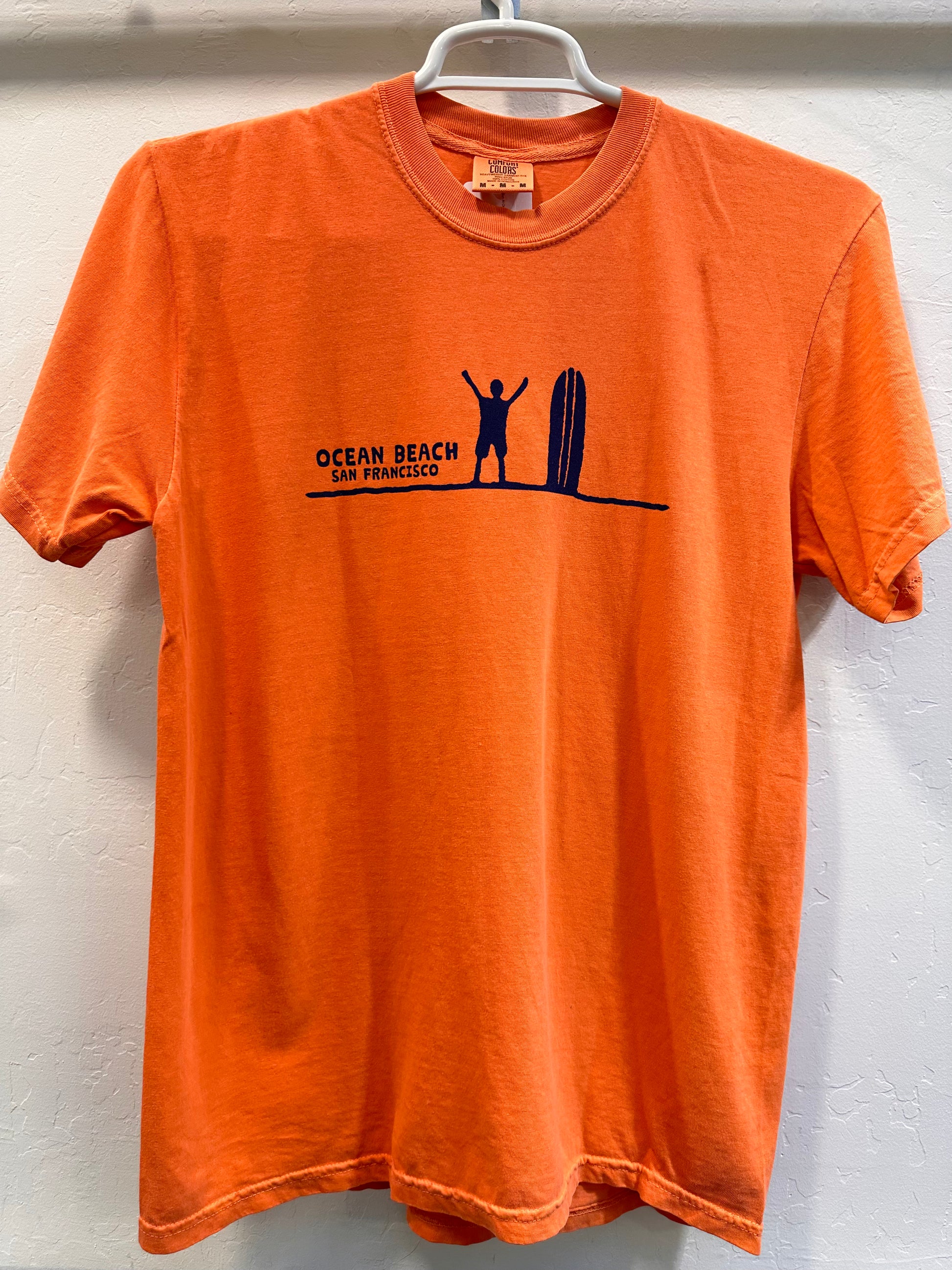 Front of a terracotta orange t-shirt displaying a minimalist navy blue graphic across the chest. It features a silhouette of a surfer standing with arms raised next to a vertical surfboard on a single horizon line, captioned with the words "OCEAN BEACH SAN FRANCISCO."