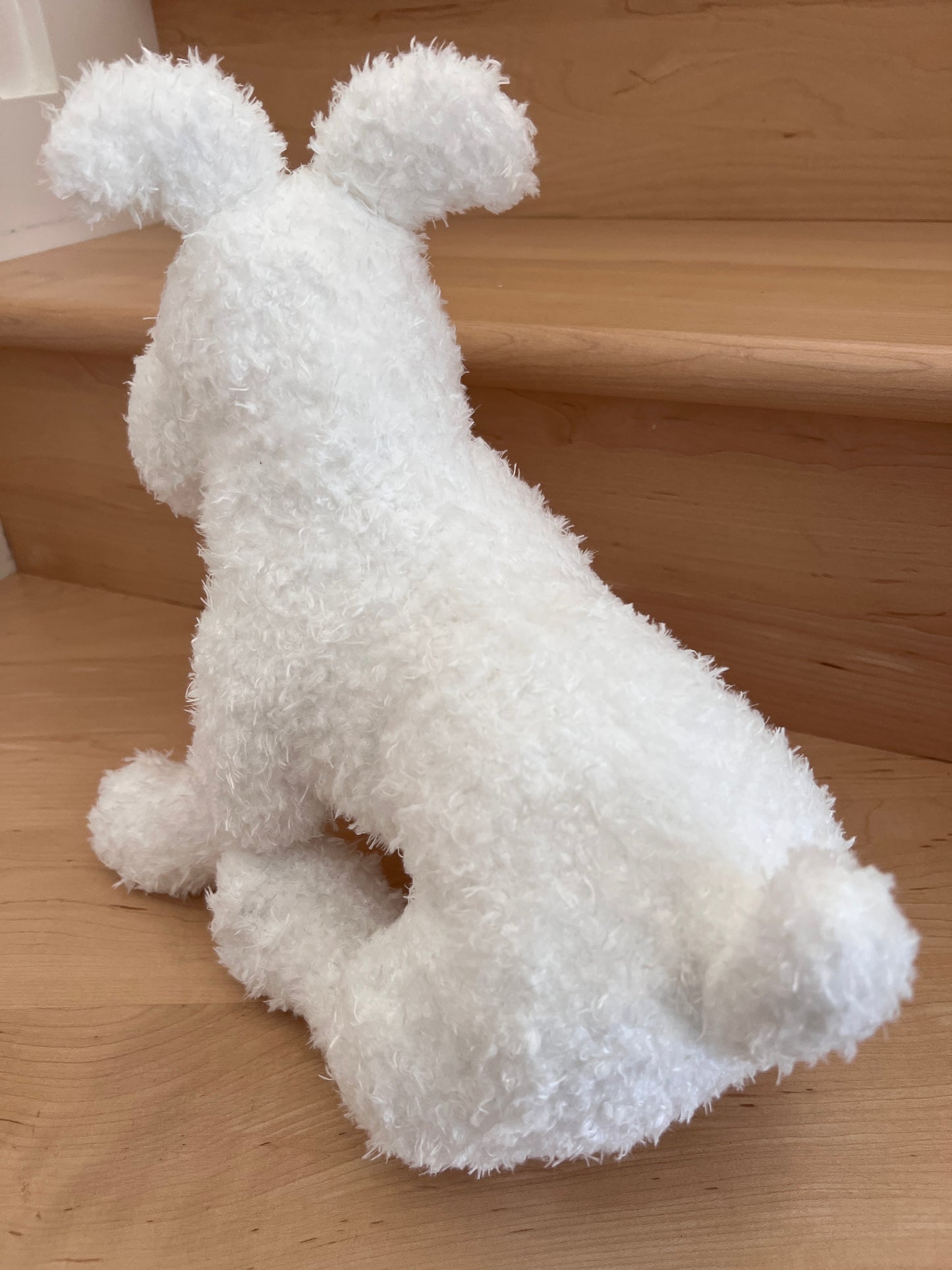 A close-up photograph showing the back and side profile of the official 14.5-inch plush Snowy toy from Tintin. The soft, fluffy white stuffed dog is sitting upright on light-colored wooden stairs, facing away and slightly to the left, highlighting its short upright tail and fluffy ears.