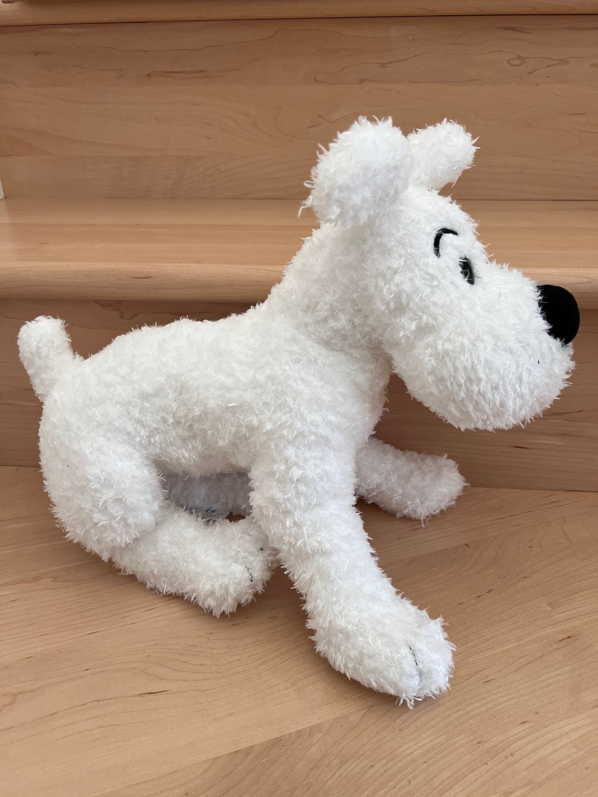 A photograph of the official 14.5-inch plush Snowy toy from Tintin, showing its side profile. The soft, bright white stuffed dog features heavy beanbag paws for easy positioning and sits upright on a light-colored wooden step against a clean background.