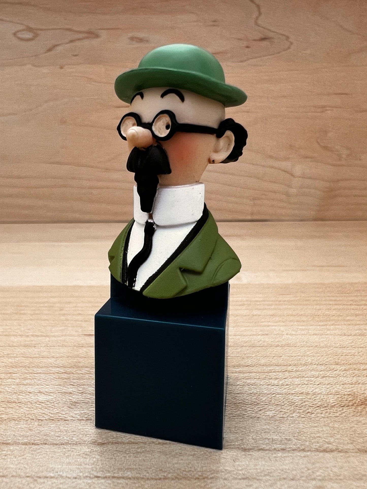 A mini PVC collectible bust figure of Professor Calculus from Tintin, featuring his green bowler hat, round glasses, and black mustache, mounted on a dark blue square pedestal.
