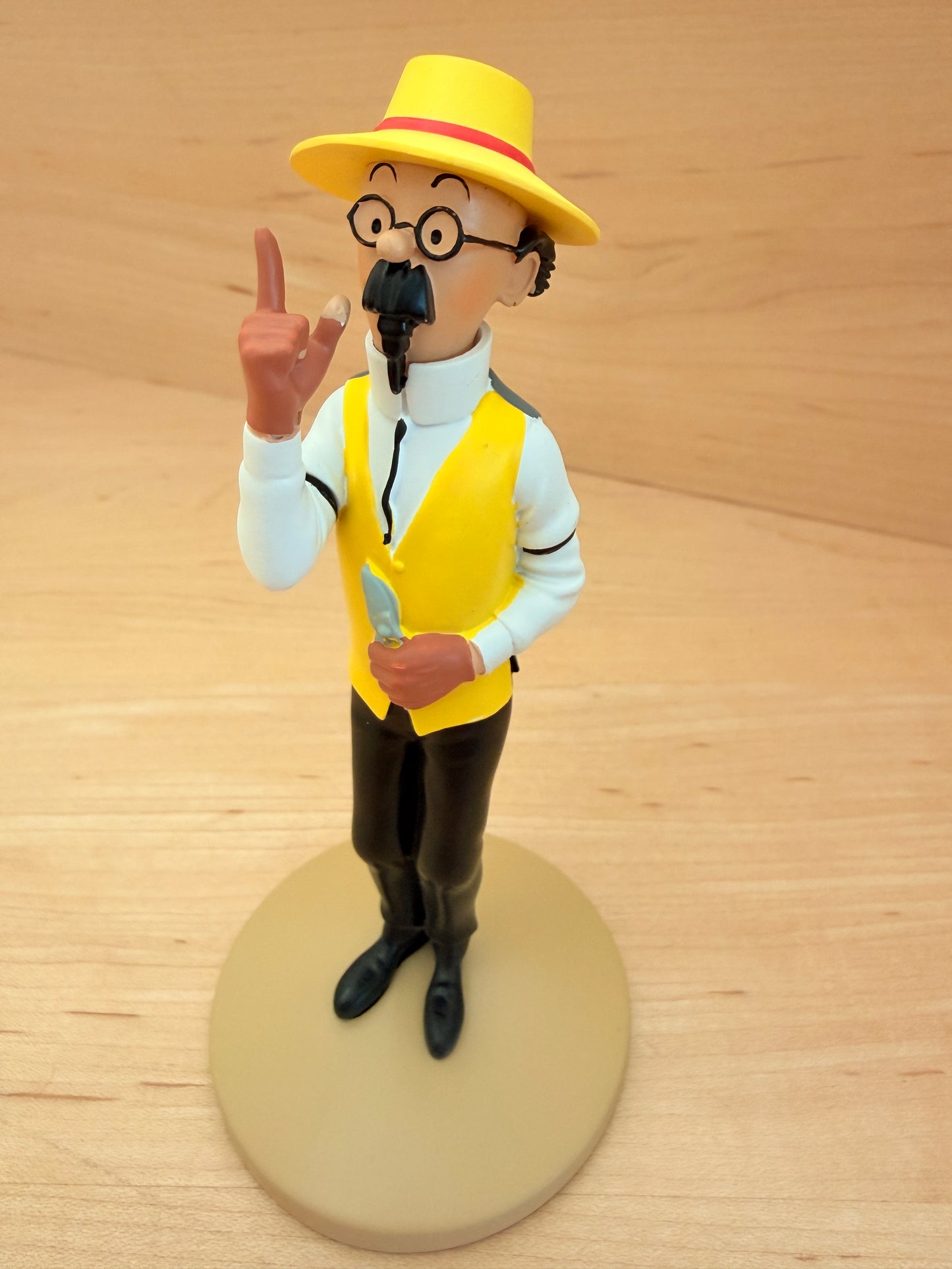 A 12 cm hand-painted resin figure of Professor Calculus from Hergé's Tintin comic The Castafiore Emerald. He is depicted standing on a round tan base, wearing a wide-brimmed yellow sun hat, a bright yellow vest over a crisp white collared shirt, black trousers, and black shoes. He holds a small silver trowel in his left hand and points upward with his right index finger as if mid-explanation.