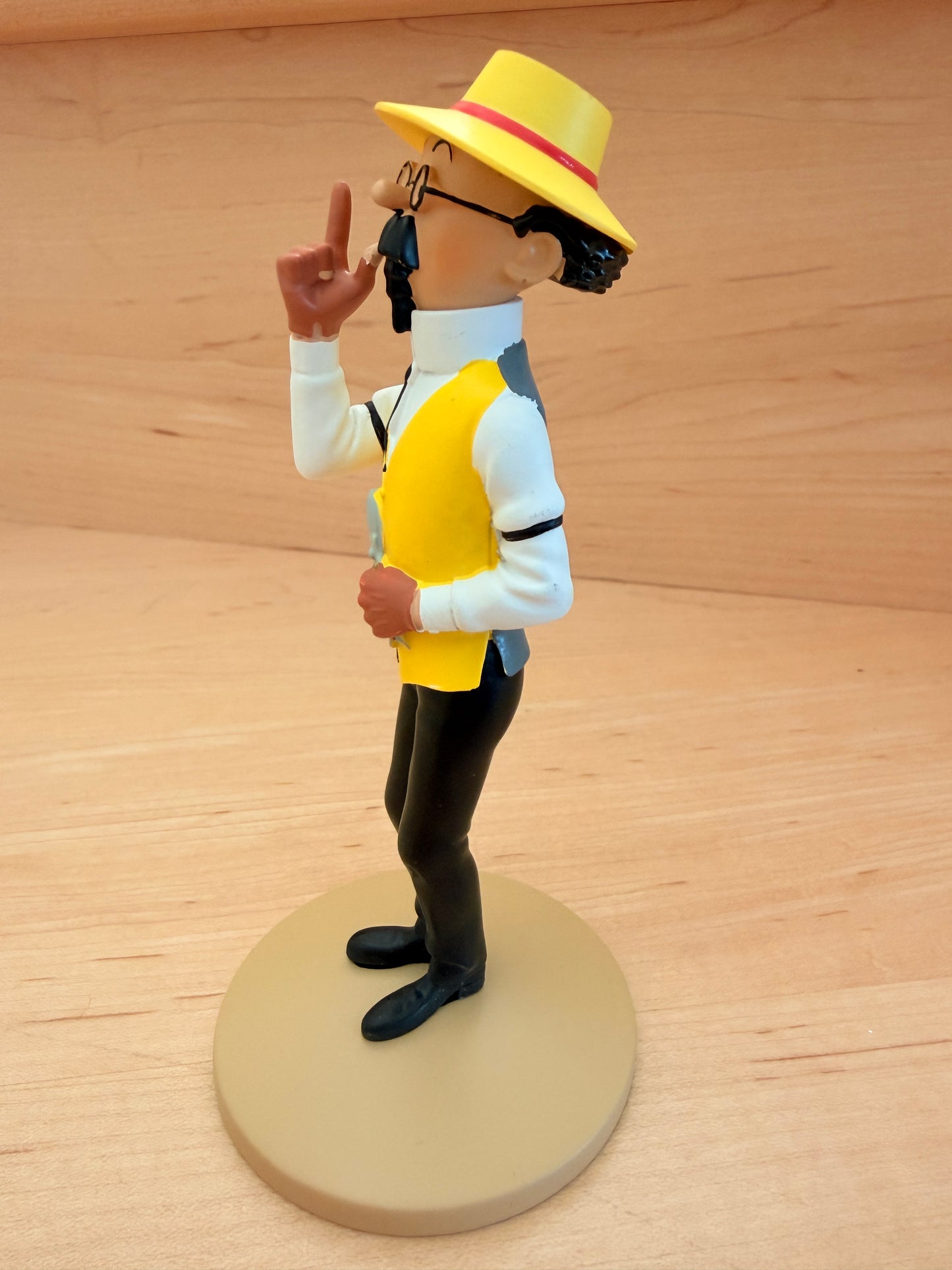 A side profile view of a 12 cm hand-painted resin figure of Professor Calculus from Tintin, showcasing his classic green coat, black trousers, and wide-brimmed yellow sun hat. He stands on a round tan base, looking to the side with his right index finger raised thoughtfully in the air as if explaining his horticultural work.