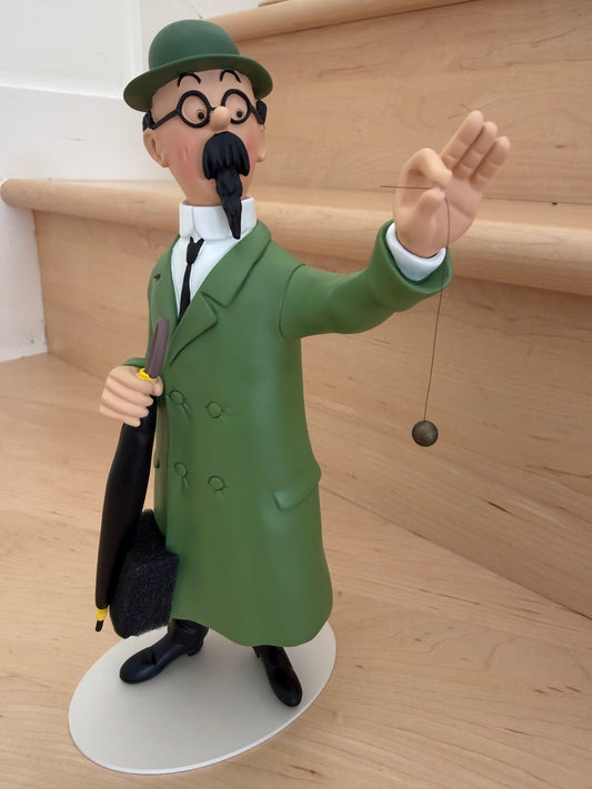 A 26cm resin collectible statue of Professor Calculus from Tintin, wearing his signature green coat and matching green bowler hat. He stands on an off-white oval base, holding a black umbrella in one hand and delicately suspending a dowsing pendulum from his raised left hand.