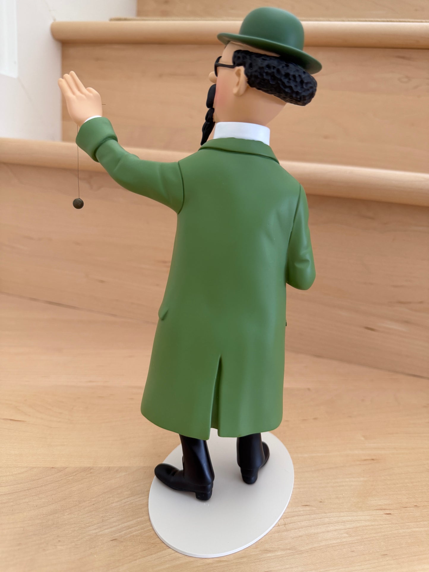 The back view of a 26cm resin collectible statue of Professor Calculus from Tintin, wearing his iconic long green coat and matching green bowler hat, standing on an off-white oval base while delicately suspending a dowsing pendulum from his raised left hand.