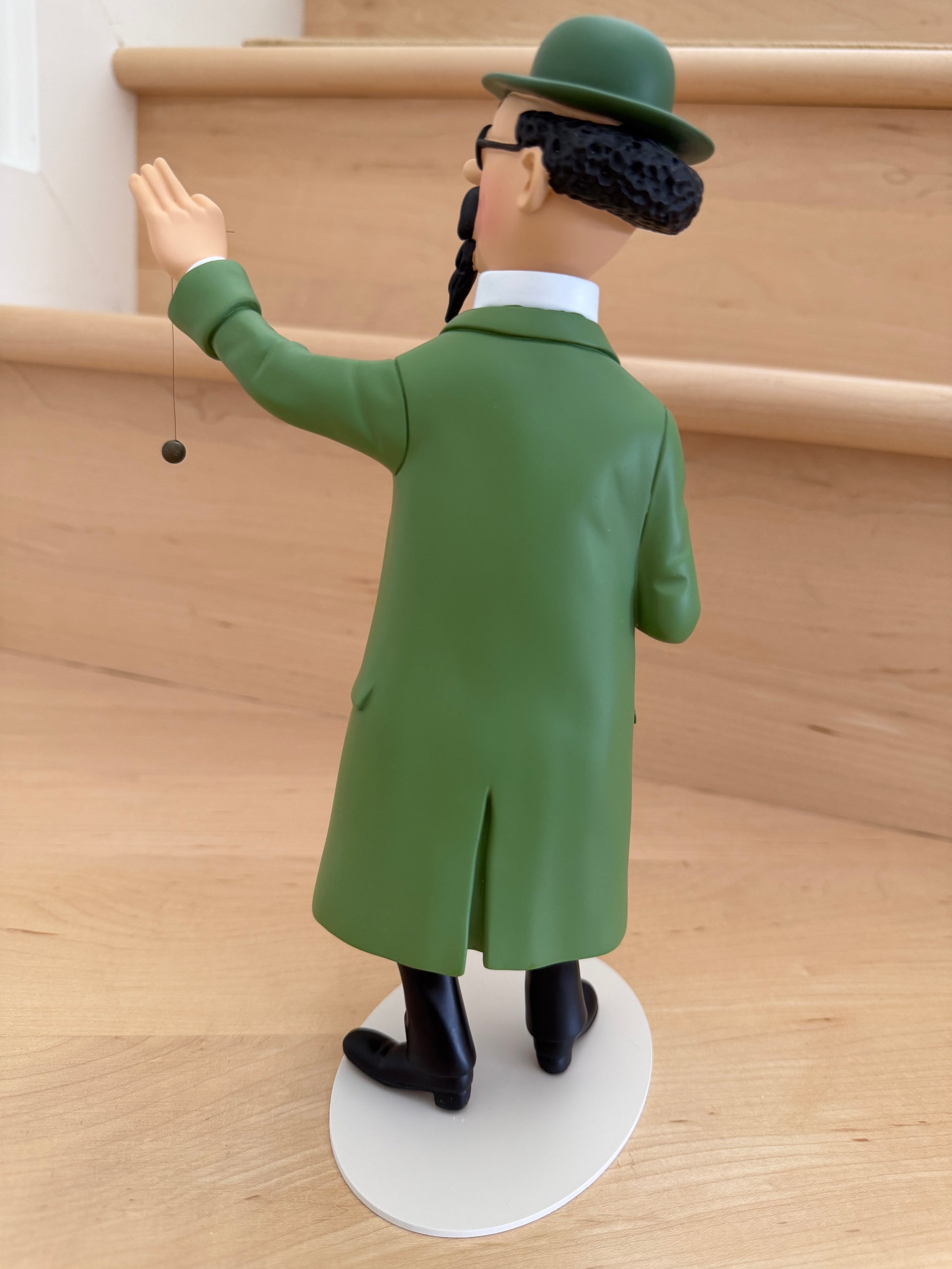 The back view of a 26cm resin collectible statue of Professor Calculus from Tintin, wearing his iconic long green coat and matching green bowler hat, standing on an off-white oval base while delicately suspending a dowsing pendulum from his raised left hand.