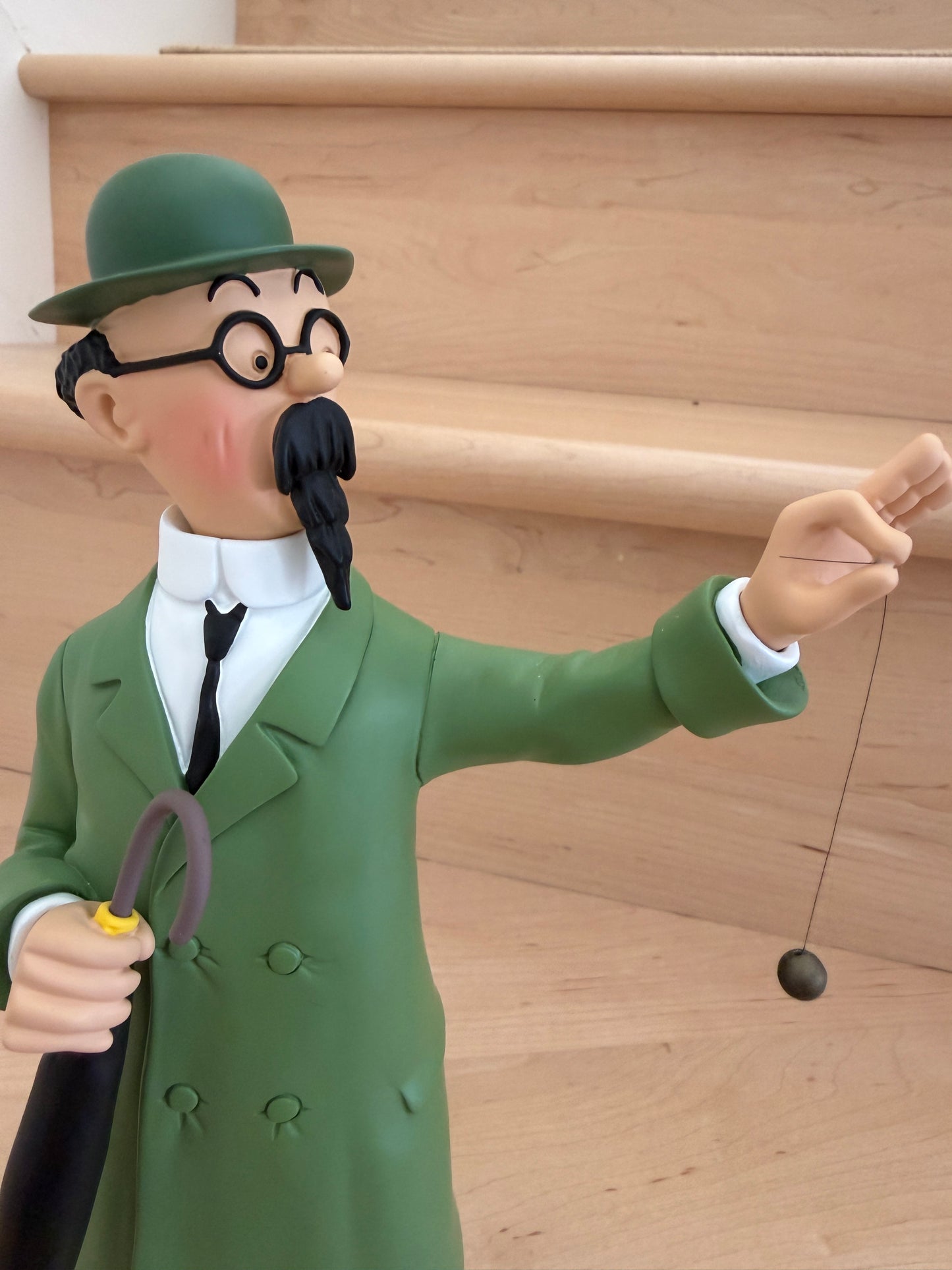 A close-up shot of a 26cm resin collectible statue of Professor Calculus from Tintin. He is depicted from the torso up wearing his classic green coat, black tie, round glasses, and green bowler hat, looking forward while holding a dark brown umbrella under his arm and balancing a small black dowsing pendulum from his extended hand.