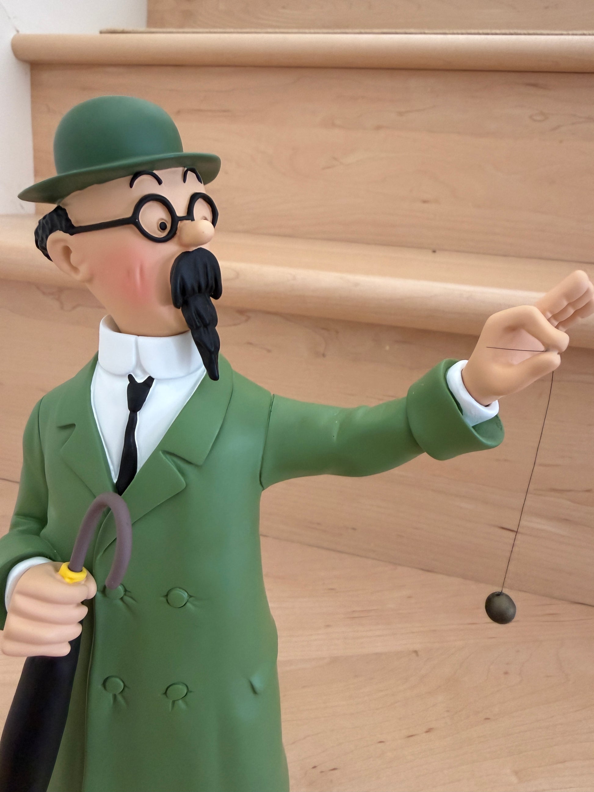 A close-up shot of a 26cm resin collectible statue of Professor Calculus from Tintin. He is depicted from the torso up wearing his classic green coat, black tie, round glasses, and green bowler hat, looking forward while holding a dark brown umbrella under his arm and balancing a small black dowsing pendulum from his extended hand.