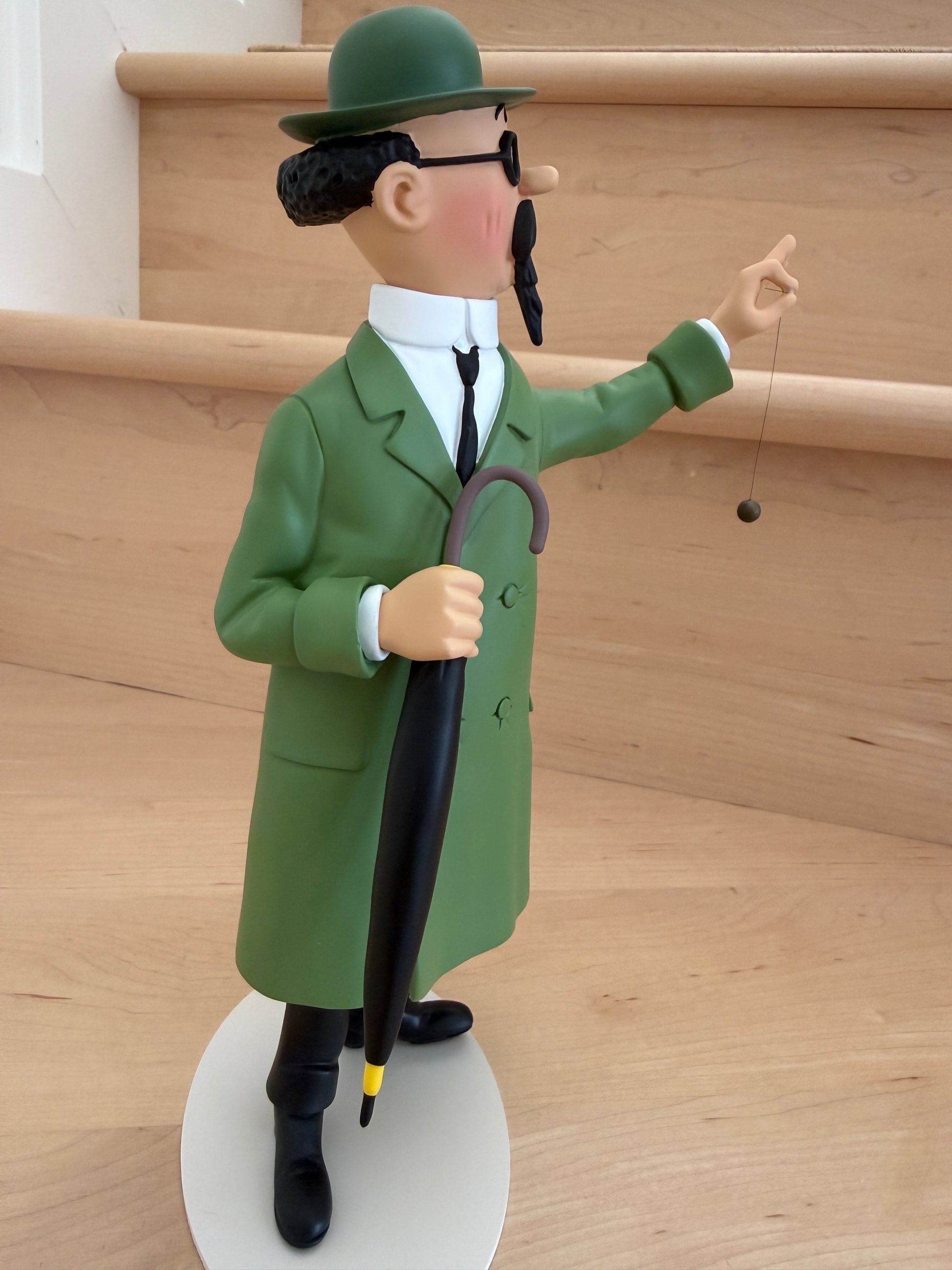 A side-profile view of a 26cm resin collectible statue of Professor Calculus from Tintin. He wears his classic long green coat, black round glasses, and green bowler hat while standing on a white oval base, looking forward with a dark brown umbrella held downward in his right hand and a small black dowsing pendulum suspended from his raised left hand.