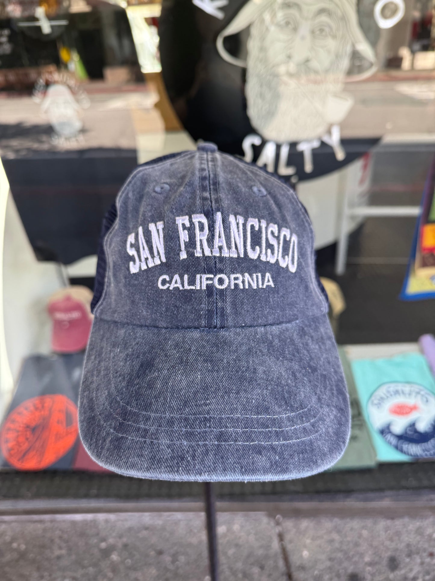 Front view of an over-washed navy blue trucker cap displayed on a stand in front of a store window. The cap features "SAN FRANCISCO" embroidered across the front in white arched capital letters with "CALIFORNIA" underneath, paired with navy mesh back panels.