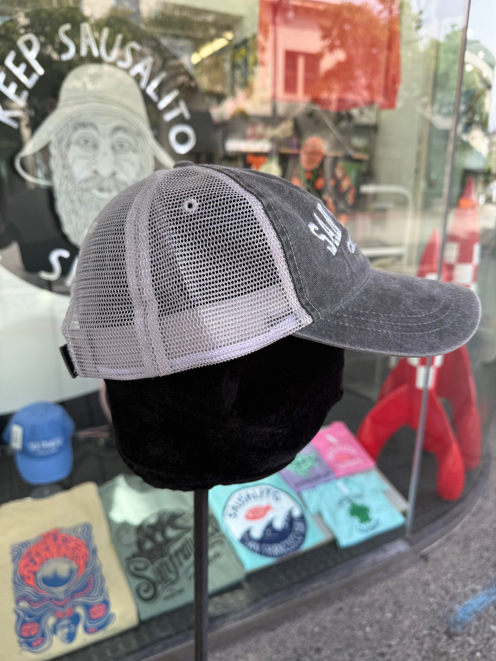 Side profile view of an over-washed charcoal gray trucker hat displayed on a mannequin head inside a store window, featuring light gray mesh back panels and white embroidered lettering visible on the front brim.