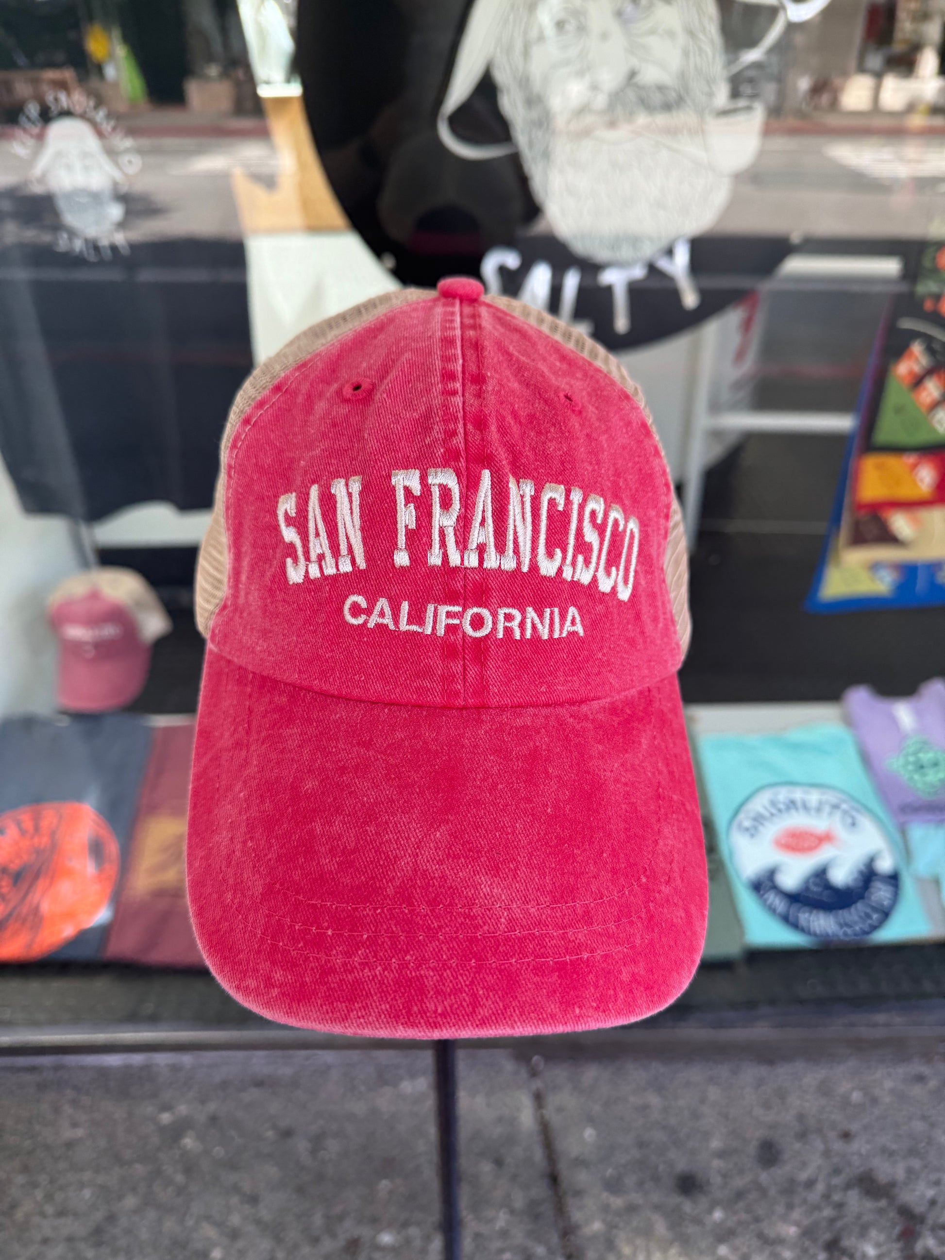 Front view of an over-washed red trucker cap displayed on a stand in front of a store window. The hat features "SAN FRANCISCO" embroidered across the front in white arched capital letters, with "CALIFORNIA" stitched below it, paired with light beige mesh back panels.