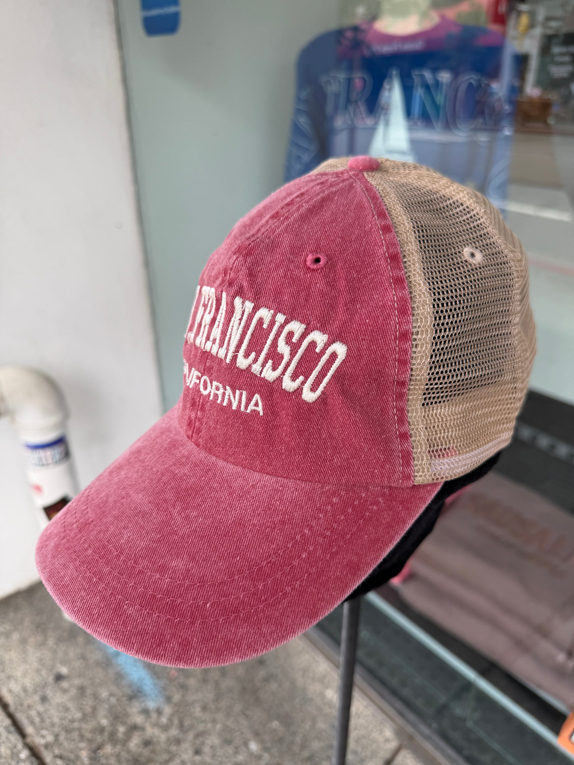 Front angle view of an over-washed wine-red trucker hat displayed on a stand in front of a store window. The cap features "SAN FRANCISCO" embroidered across the front in white arched capital letters, with "CALIFORNIA" stitched below it, paired with light beige mesh back panels.