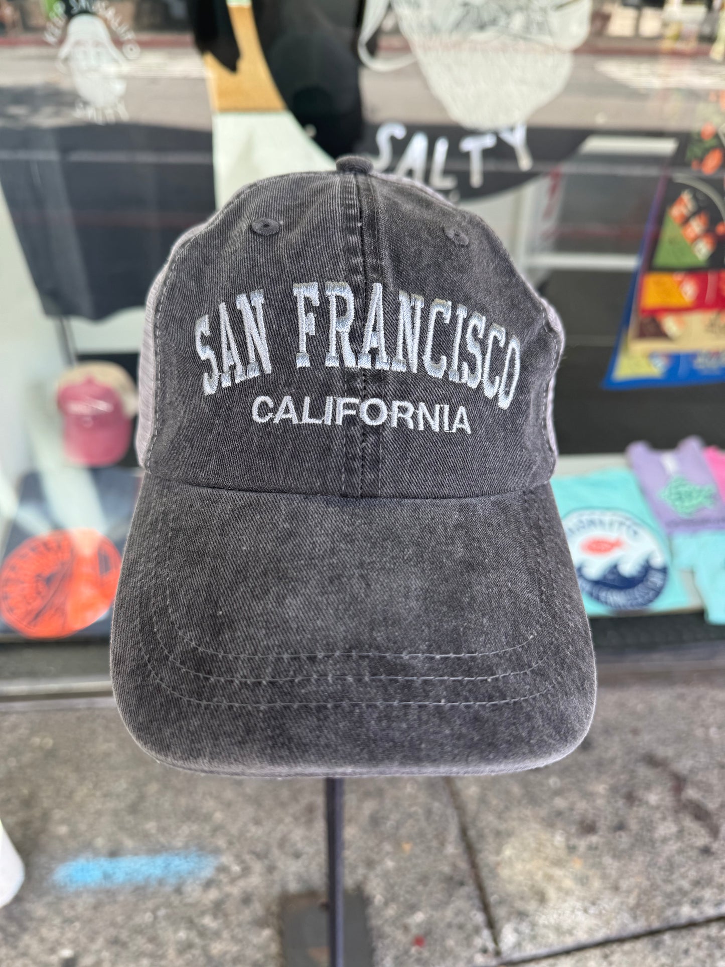 Front view of an over-washed black trucker hat displayed on a stand in front of a store window. The cap features "SAN FRANCISCO" embroidered across the front in white arched capital letters, with "CALIFORNIA" stitched directly underneath, paired with dark gray mesh back panels.
