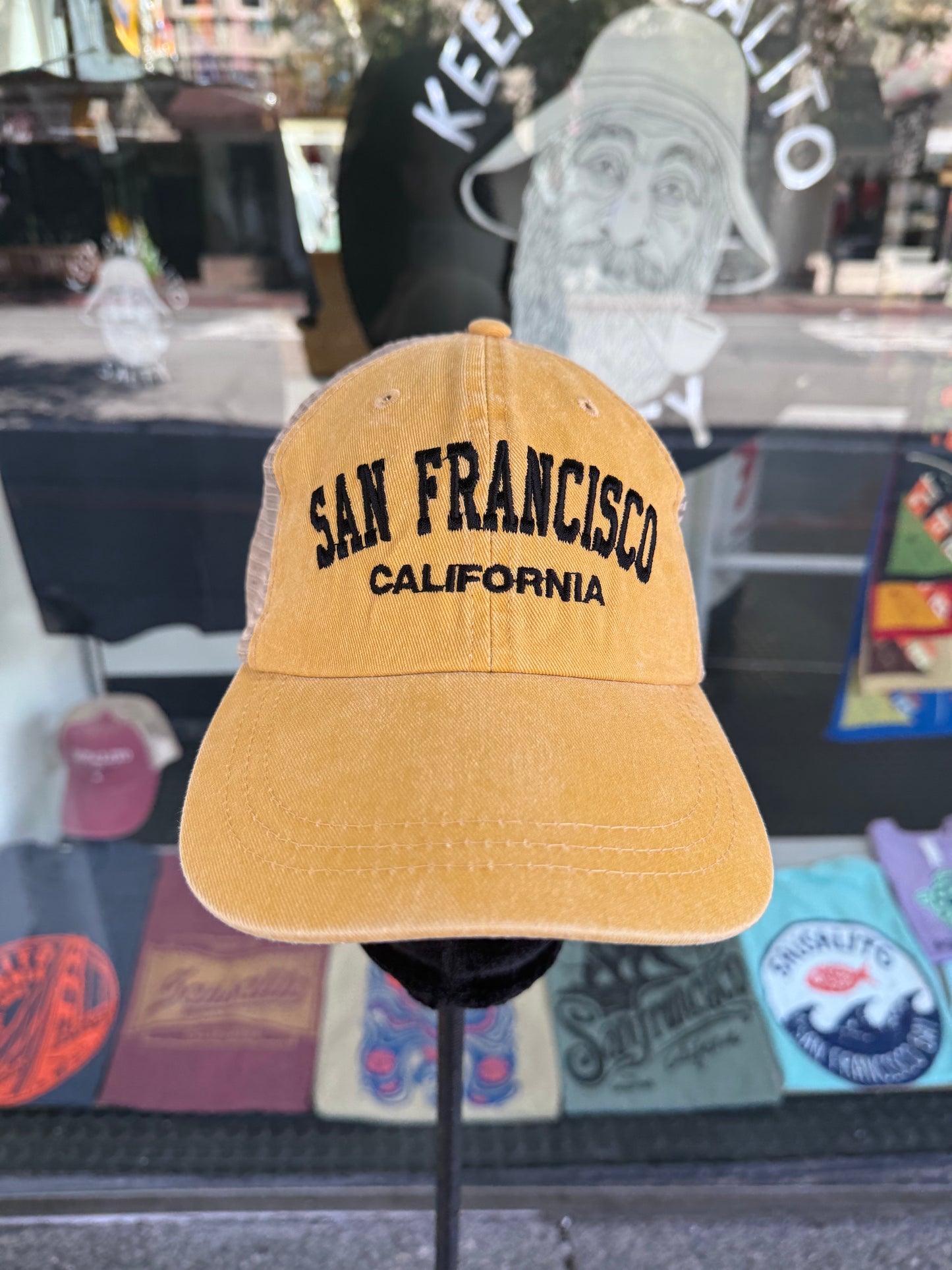 Front view of an over-washed mango yellow trucker hat displayed on a stand in front of a store window. The cap features "SAN FRANCISCO" embroidered across the front in dark navy arched capital letters, with "CALIFORNIA" stitched directly underneath, paired with light tan mesh back panels.