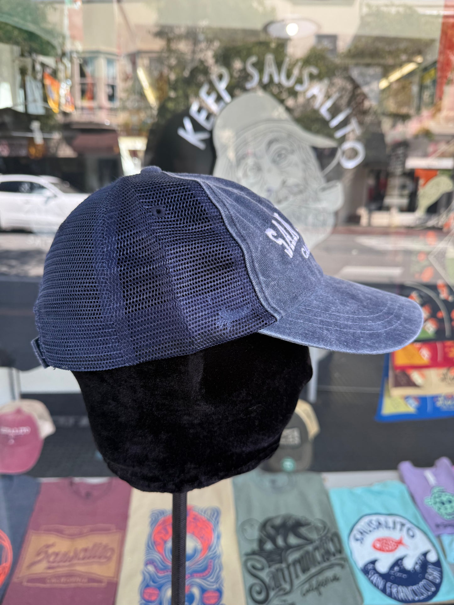 Side profile view of an over-washed navy blue trucker hat displayed on a mannequin head inside a store window, featuring dark navy mesh side panels and white embroidered lettering visible on the front brim.