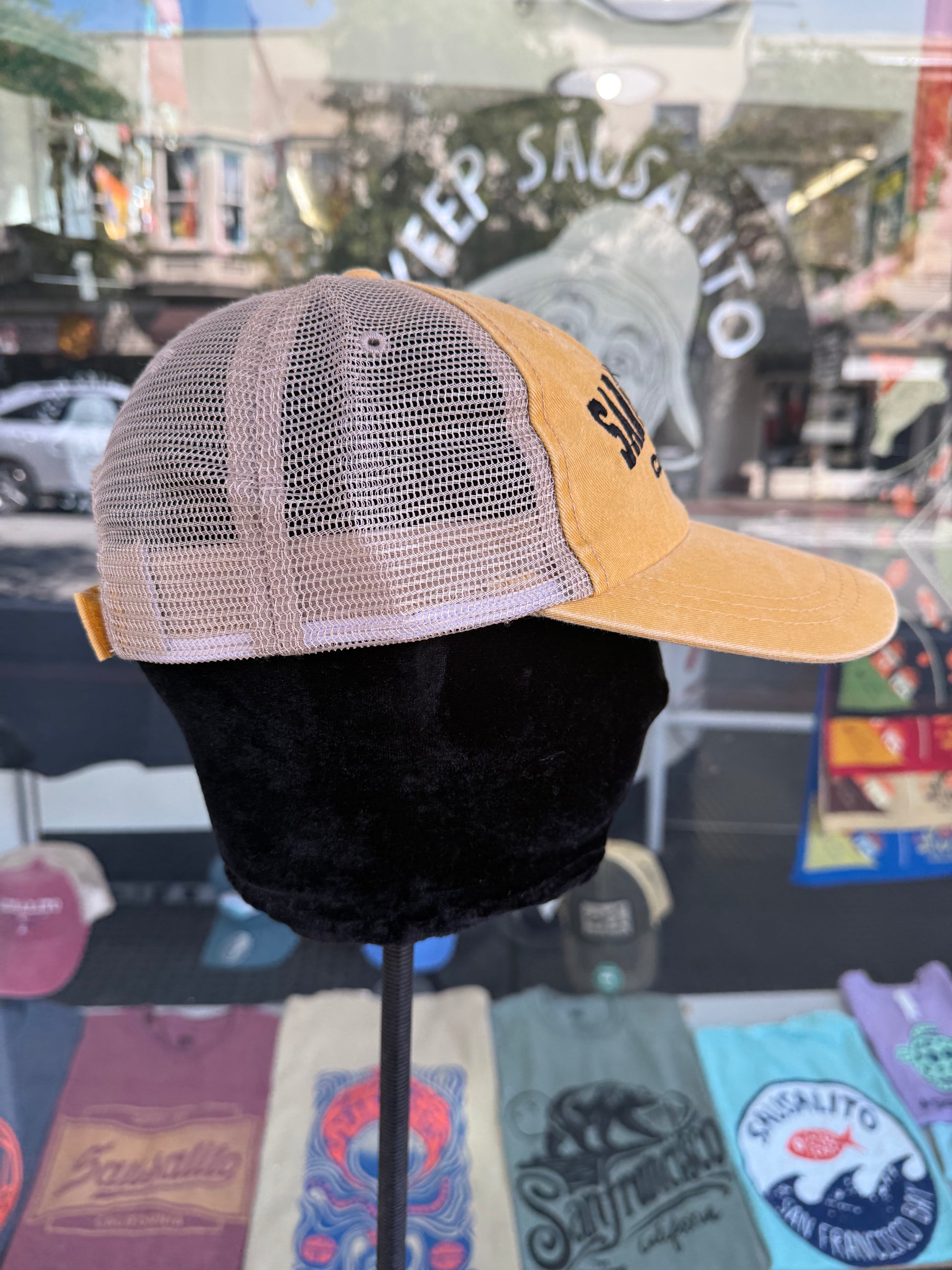 Side profile view of an over-washed mango yellow trucker hat displayed on a mannequin head inside a store window, featuring light tan mesh side panels and dark navy embroidered lettering visible on the front brim.