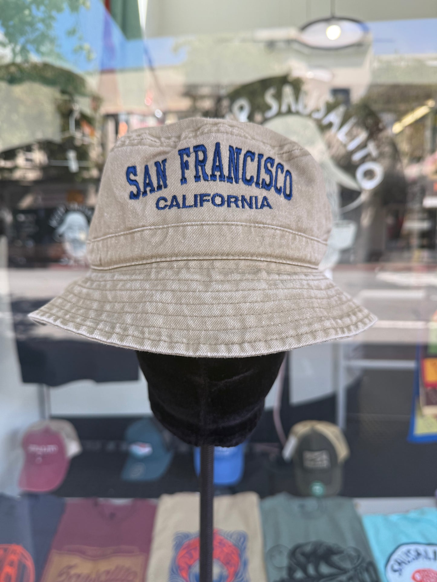 A front view of a beige, over-washed 100% cotton bucket hat displayed on a black mannequin stand in front of a store window. The front features the words "SAN FRANCISCO" cleanly embroidered in blue block letters arched over "CALIFORNIA" in smaller blue text. The reflection in the window shows the storefront lettering "Sausalito Ferry Co." and an indoor light fixture.