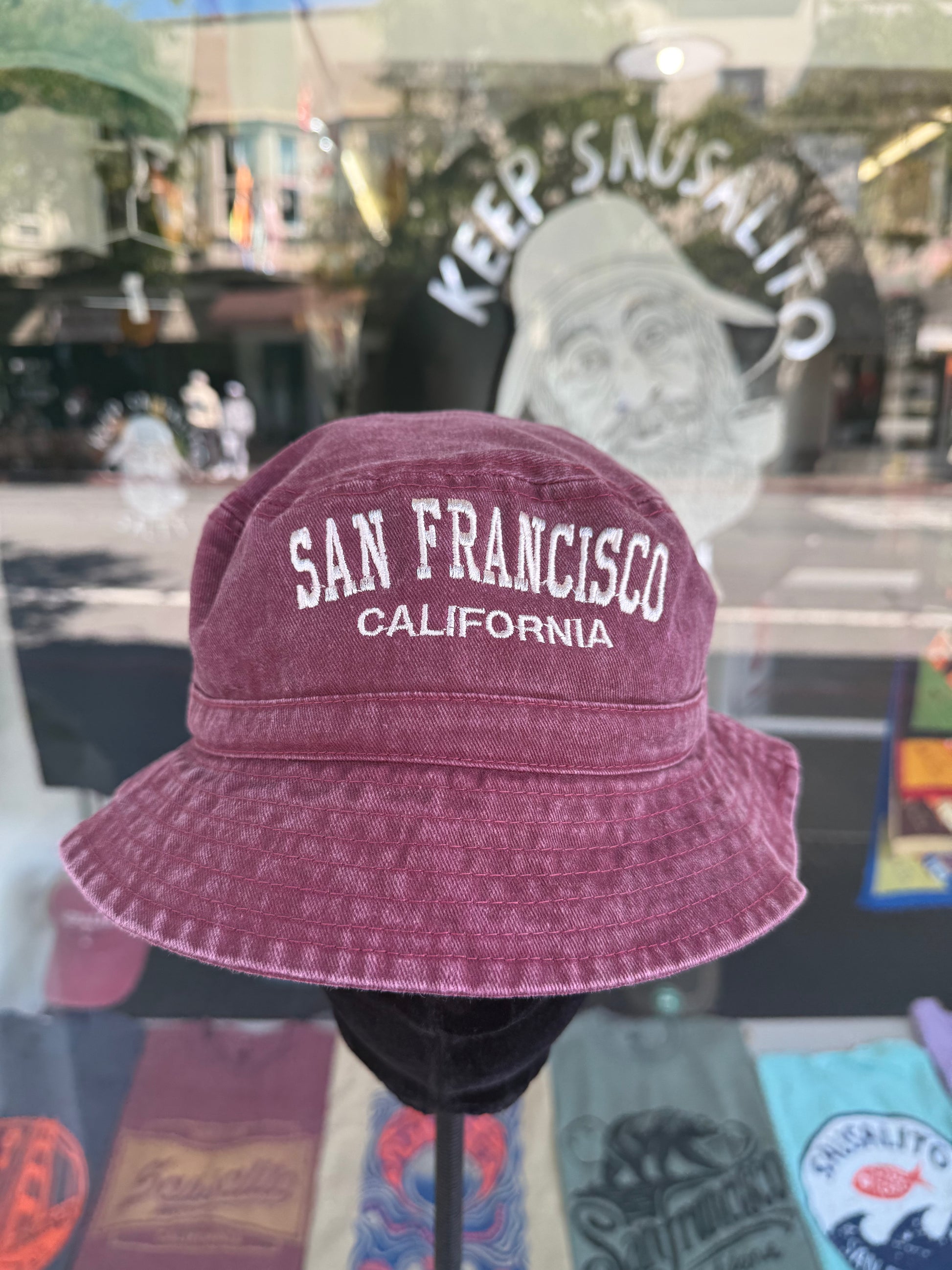 A front view of a burgundy, over-washed 100% cotton bucket hat displayed on a black mannequin stand in front of a store window. The front features the words "SAN FRANCISCO" cleanly embroidered in white block letters arched over "CALIFORNIA" in smaller white text. The reflection in the window shows the storefront sign "KEEP SAUSALITO SALTY" and a portrait illustration.