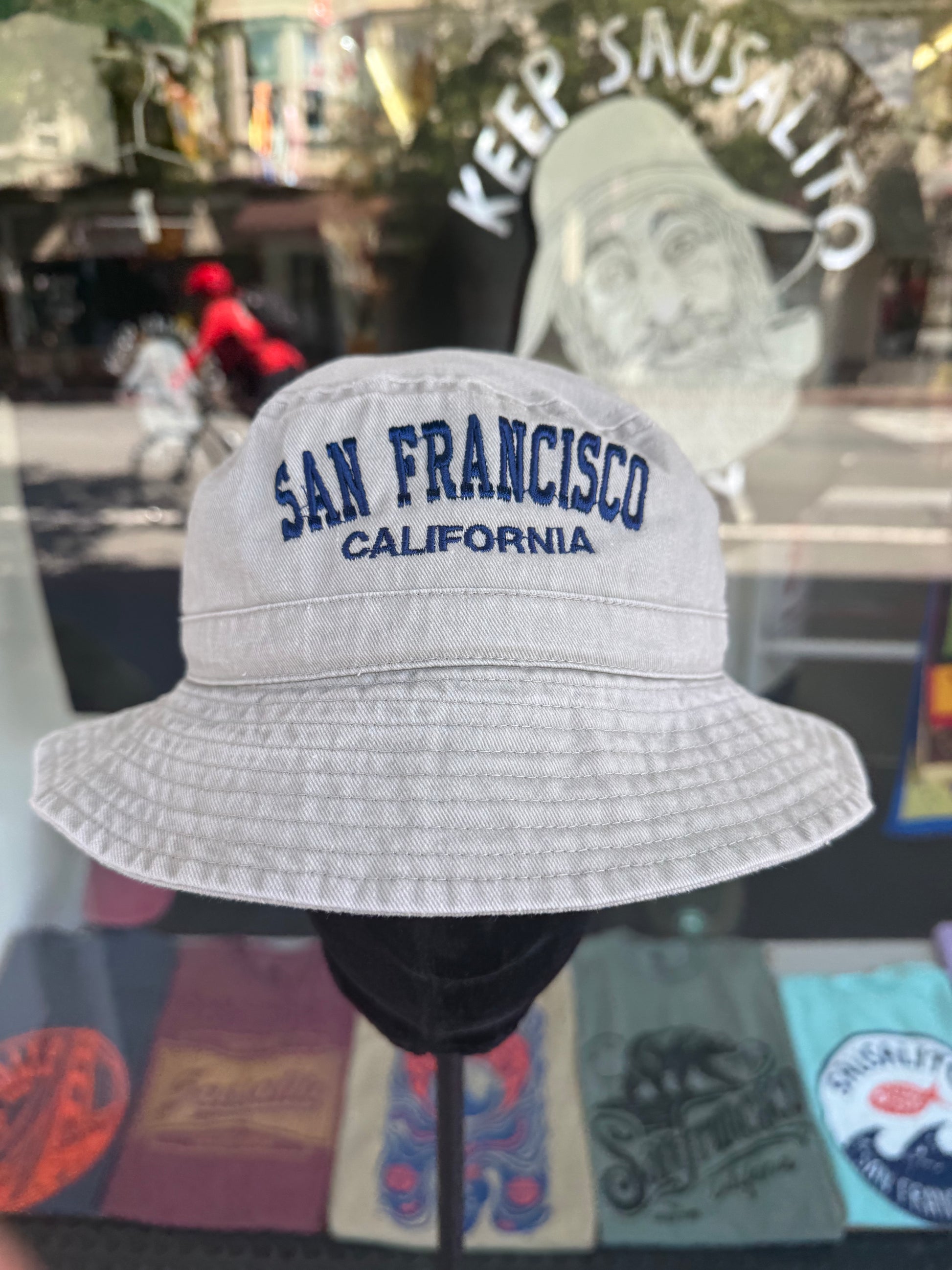 A front view of a khaki beige, over-washed 100% cotton bucket hat displayed on a mannequin stand in front of a store window. The front panel features the text "SAN FRANCISCO" cleanly embroidered in white block letters arched over "CALIFORNIA" in smaller white script.