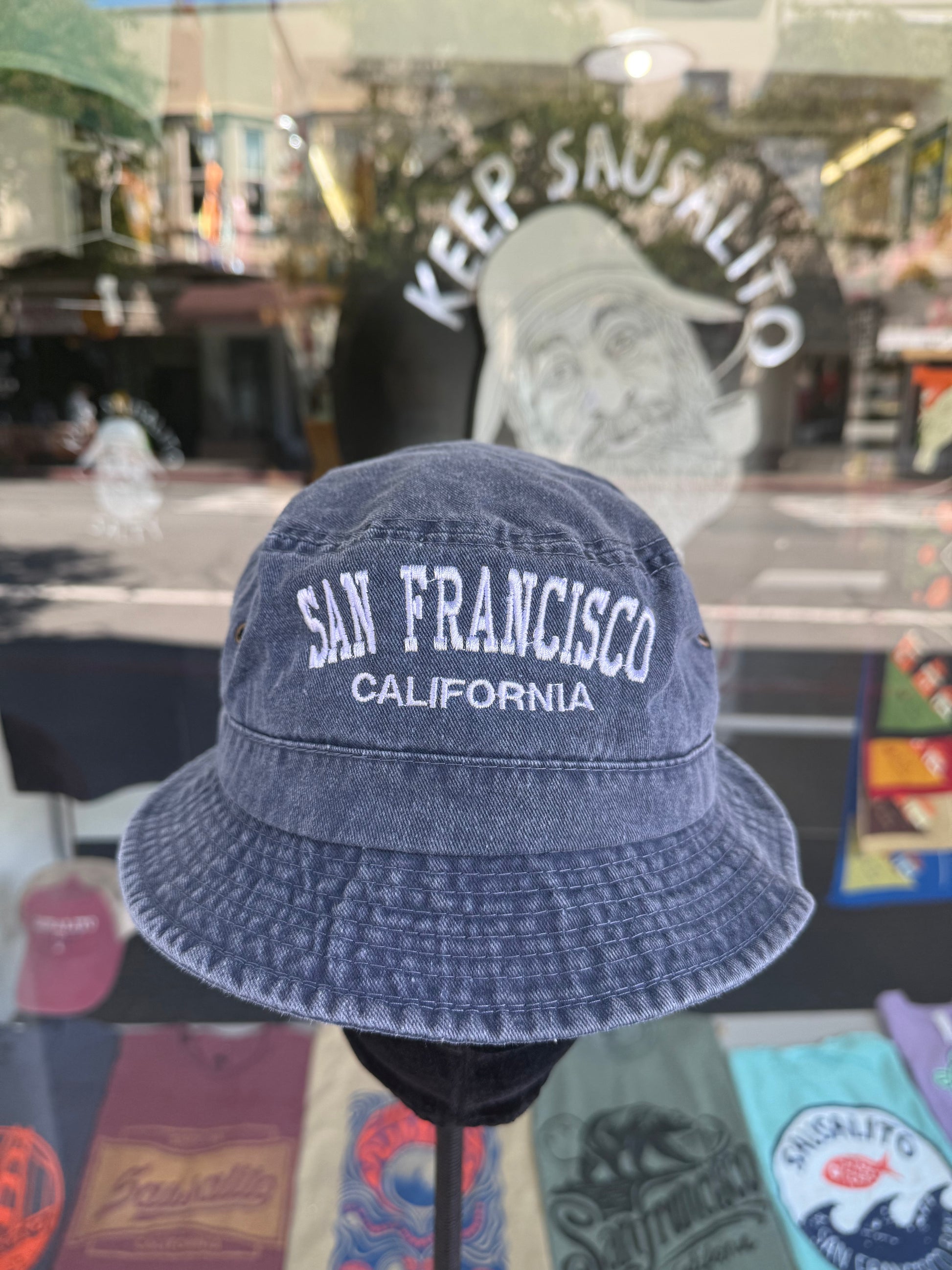 A front view of a navy blue, over-washed 100% cotton bucket hat displayed on a black mannequin stand in front of a store window. The front features the words "SAN FRANCISCO" cleanly embroidered in white block letters arched over "CALIFORNIA" in smaller white text. The reflection in the window shows the storefront sign "KEEP SAUSALITO SALTY" and a portrait illustration.