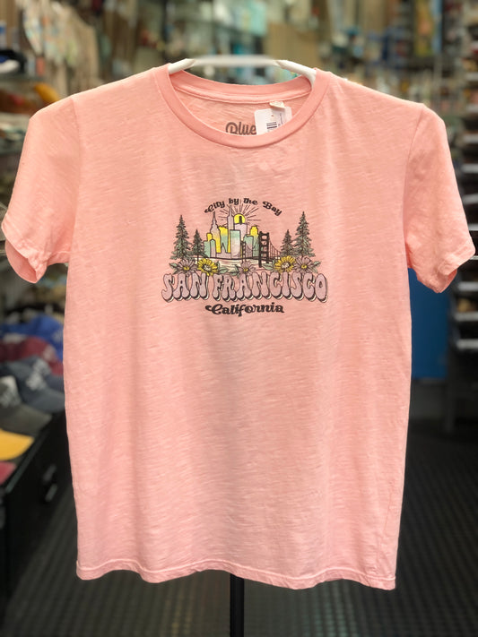 A pink women's t-shirt featuring a colorful vintage-inspired "San Francisco California" graphic illustration with the Golden Gate Bridge, city skyline, and pine trees, displayed on a hanger.