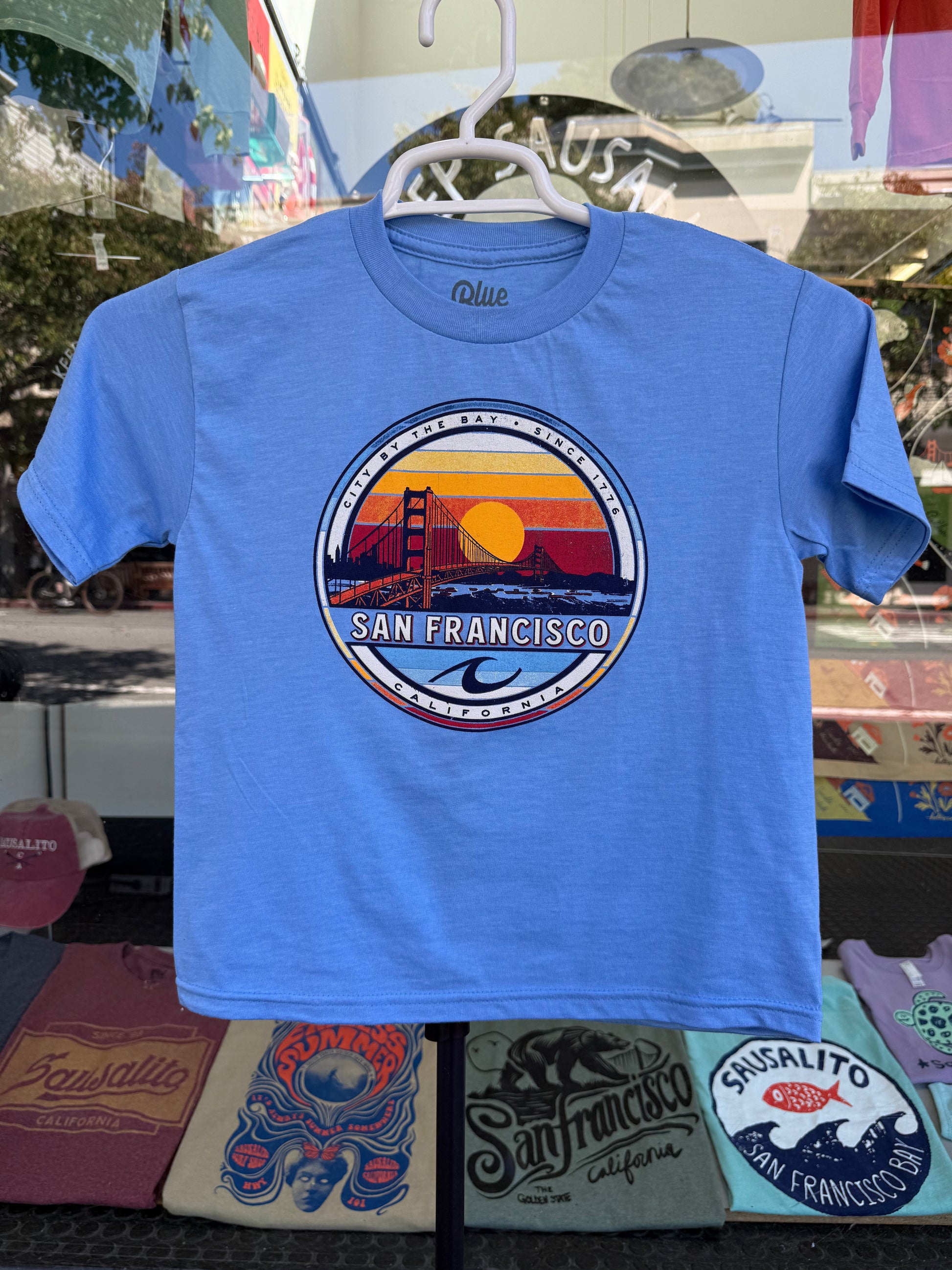 A pacific blue kids' t-shirt featuring a circular retro graphic emblem with a sunset over the Golden Gate Bridge and a blue ocean wave, with 'San Francisco California' text, displayed on a white hanger outside a clothing shop.