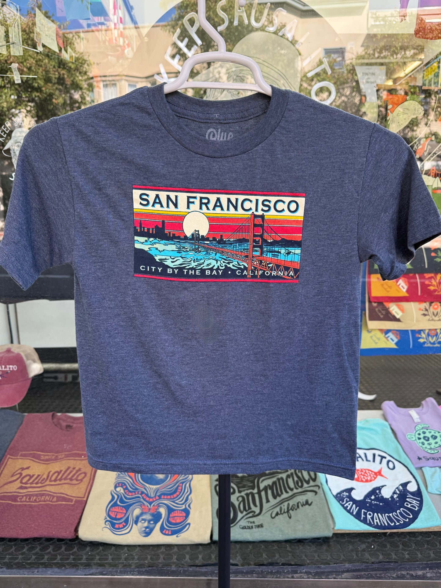 Blue t-shirt with San Francisco name drop against sunset design on a hanger photographed outside a storefront. 