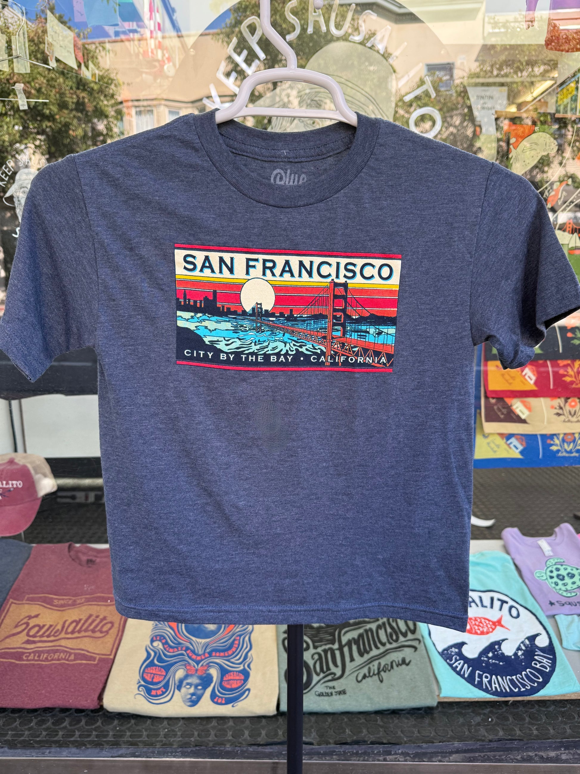 Blue t-shirt with San Francisco name drop against sunset design on a hanger photographed outside a storefront. 