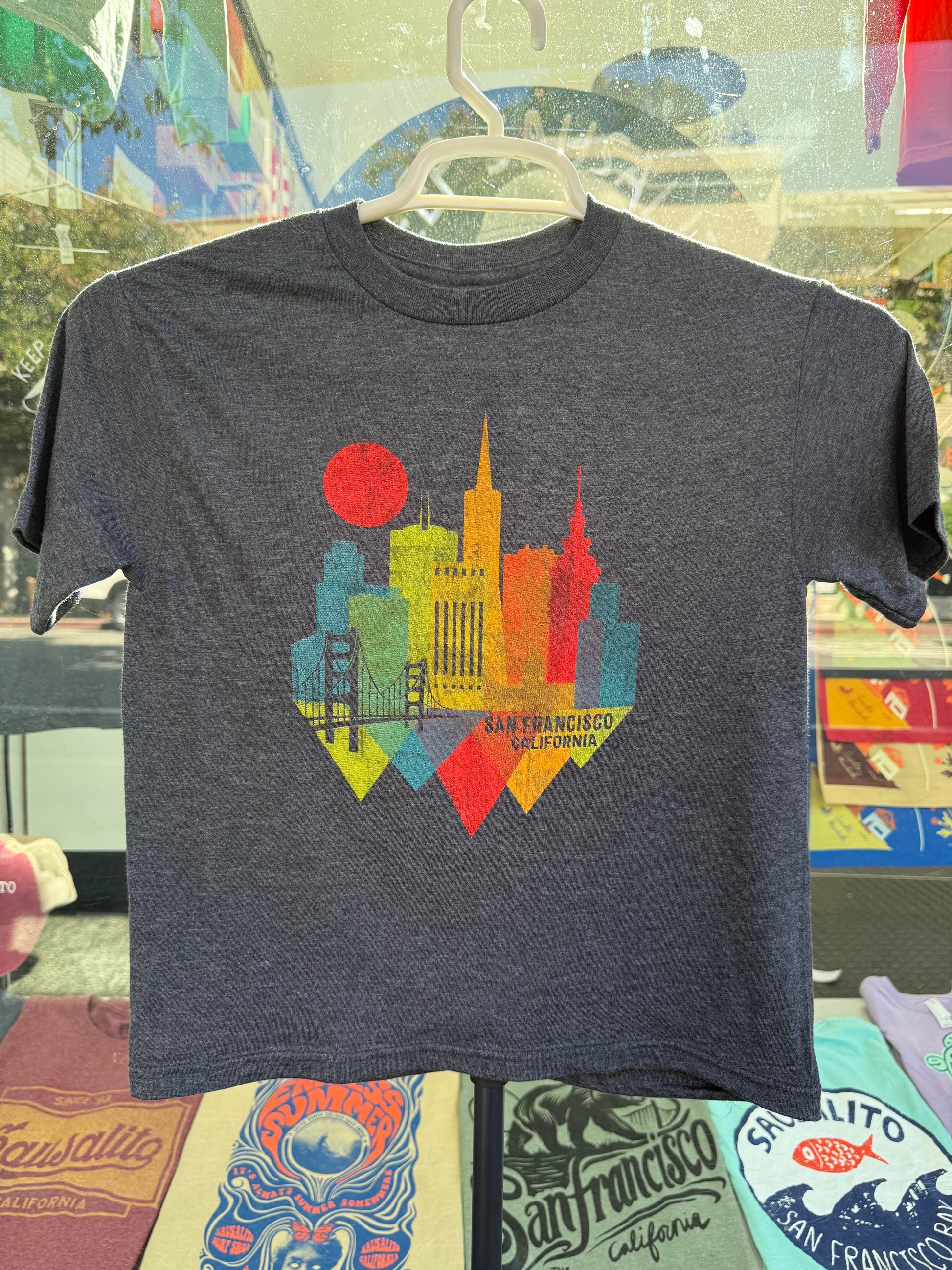 A dark gray heathered kids' t-shirt featuring a colorful geometric shard graphic illustration of the San Francisco skyline, city buildings, and a red sun, displayed on a white hanger outside a shop.