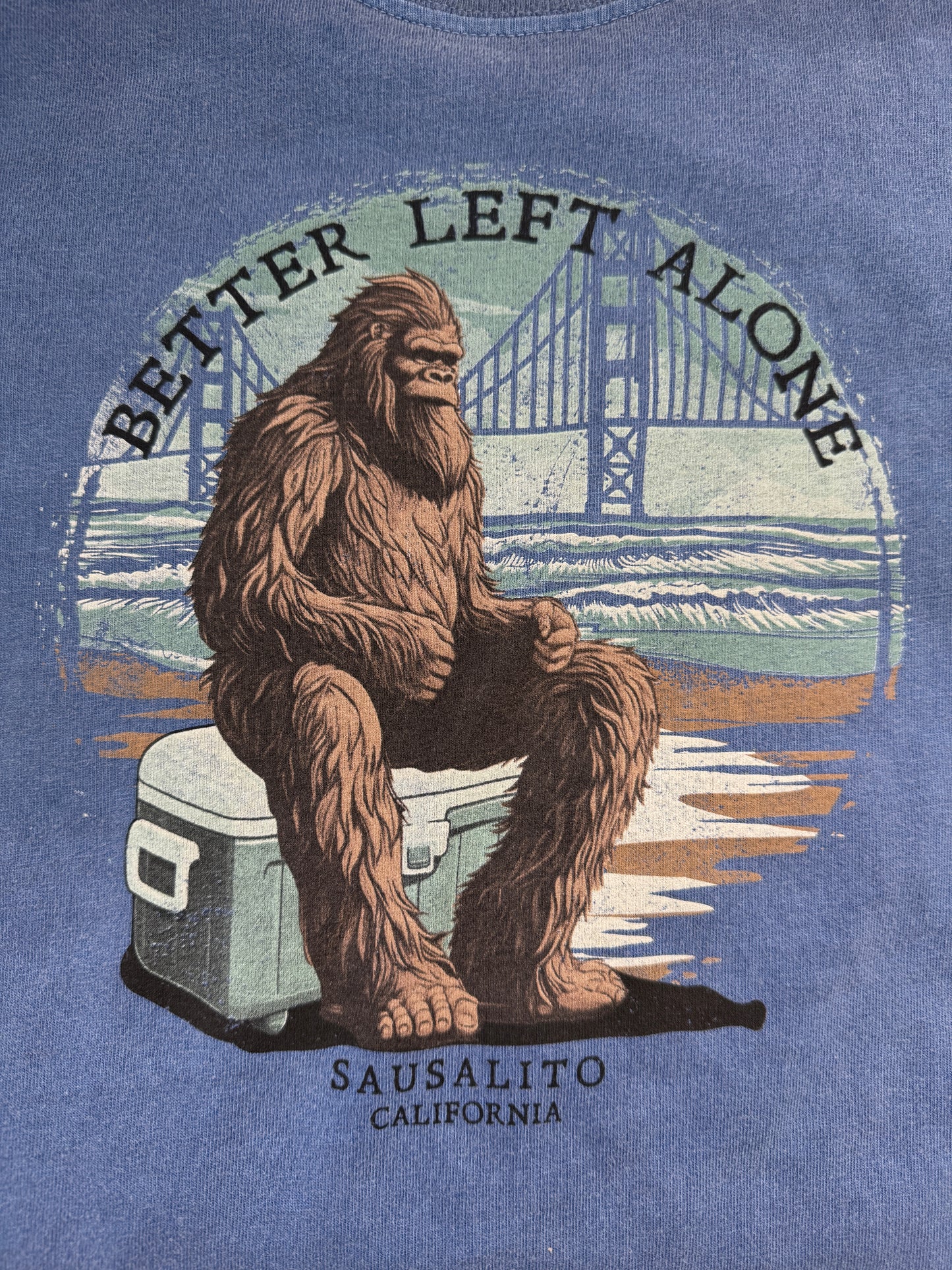 A close-up view of the circular graphic on the blue pigment-dyed t-shirt. It features a detailed illustration of a hairy Sasquatch sitting grumpily with crossed arms on a white and blue cooler at the beach. The Golden Gate Bridge spans the background behind him, framed by the arched text "BETTER LEFT ALONE" at the top and "SAUSALITO CALIFORNIA" at the bottom.