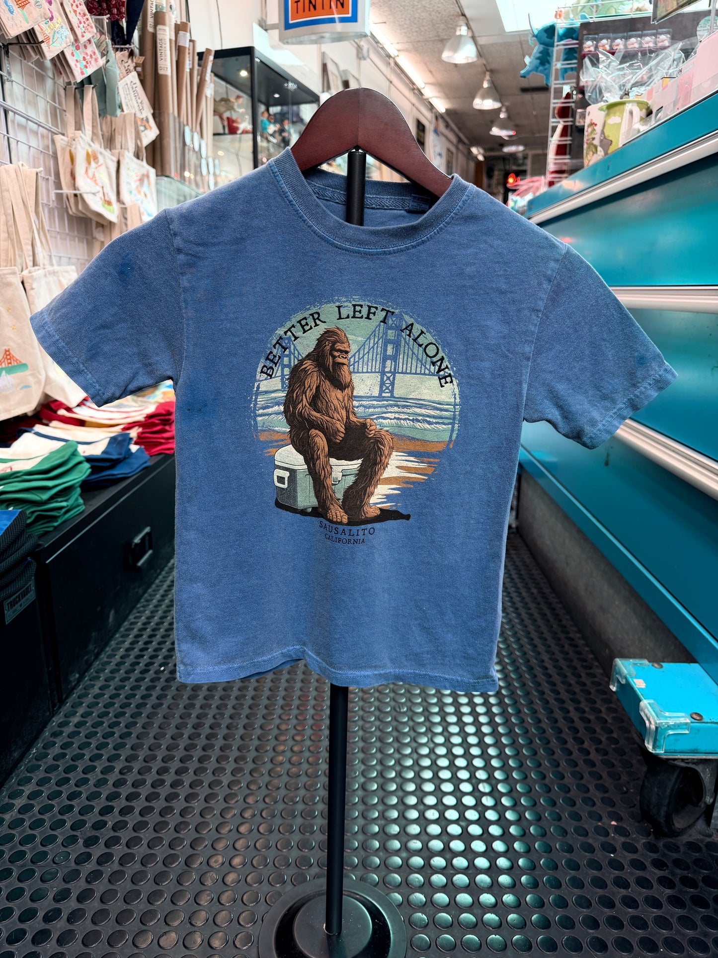 A front view of a blue, pigment-dyed short-sleeve kid's t-shirt hanging on a wooden hanger in a retail store. The circular graphic on the chest features a hairy Sasquatch sitting grumpily with arms crossed on a cooler at the beach, framed by the Golden Gate Bridge in the background under the text "BETTER LEFT ALONE."