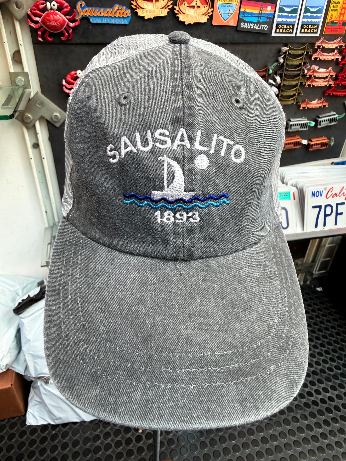 A front view of a dark grey, pigment-dyed unstructured trucker hat with a light grey mesh back. The front panel features white embroidered block lettering that reads "SAUSALITO" arched over a white and blue embroidered sailboat on waves with a full moon, and the year "1893" stitched below.