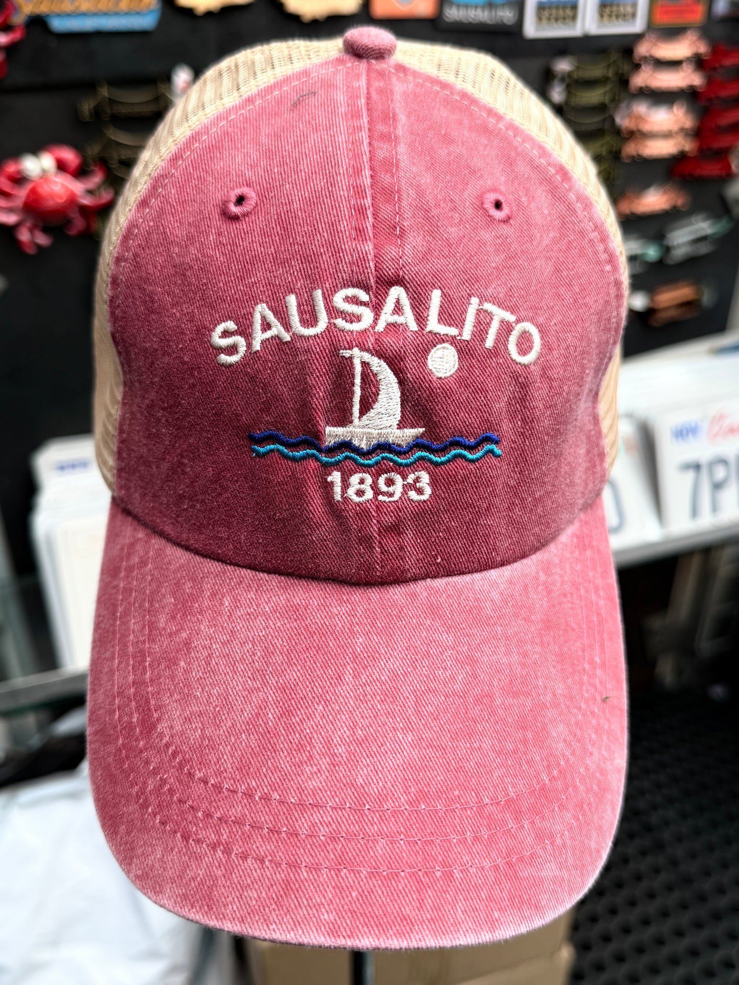 A close-up front view of a maroon, pigment-dyed unstructured trucker hat with a tan mesh back. The front features white embroidered block lettering that reads "SAUSALITO" arched over a white and blue embroidered sailboat on waves with a full moon, and the year "1893"