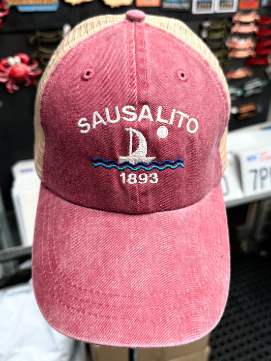 A close-up front view of a maroon, pigment-dyed unstructured trucker hat with a tan mesh back. The front features white embroidered block lettering that reads "SAUSALITO" arched over a white and blue embroidered sailboat on waves with a full moon, and the year "1893"