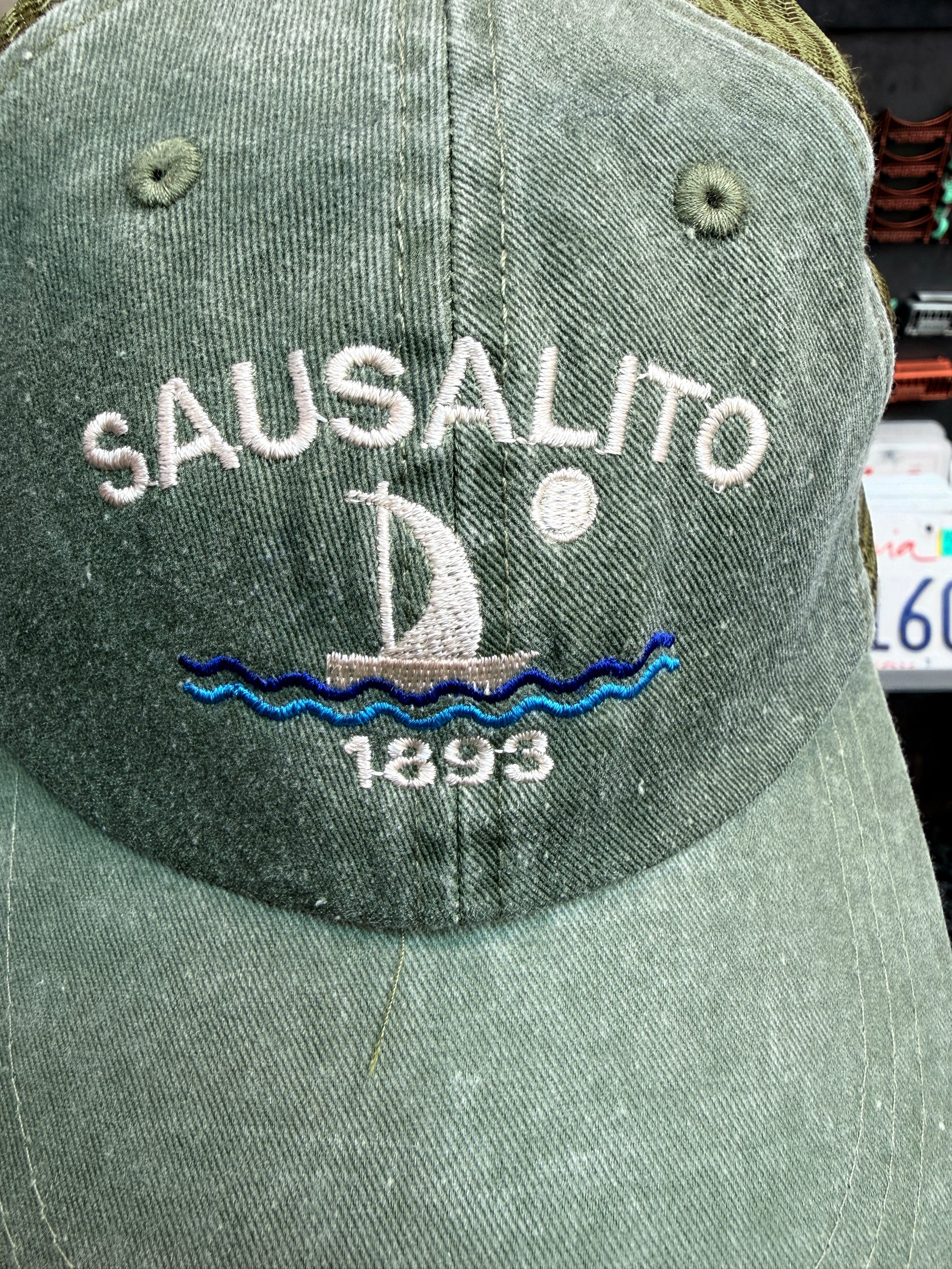 A close-up front view of the sage green, pigment-dyed unstructured trucker hat, highlighting the crisp white embroidered block lettering of "SAUSALITO" curved over a classic sailboat design on blue stitched waves beneath a small white full moon, with the year "1893" cleanly embroidered below.