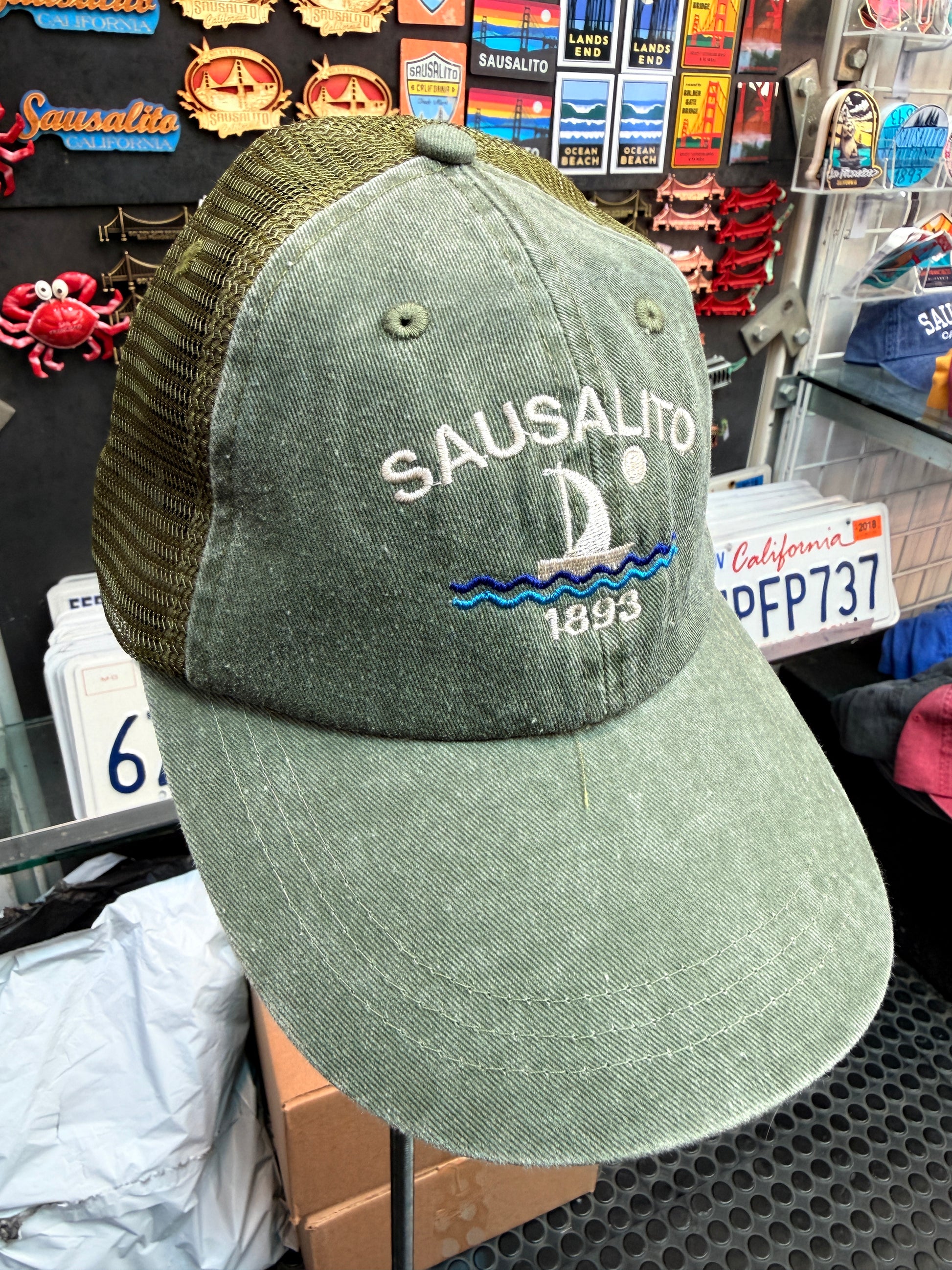 A close-up front view of an sage green, pigment-dyed unstructured trucker hat with a matching olive green mesh back. The front panel features white embroidered block lettering that reads "SAUSALITO" arched over a white and blue embroidered sailboat sailing on blue waves beneath a full moon, with the year "1893" stitched below.