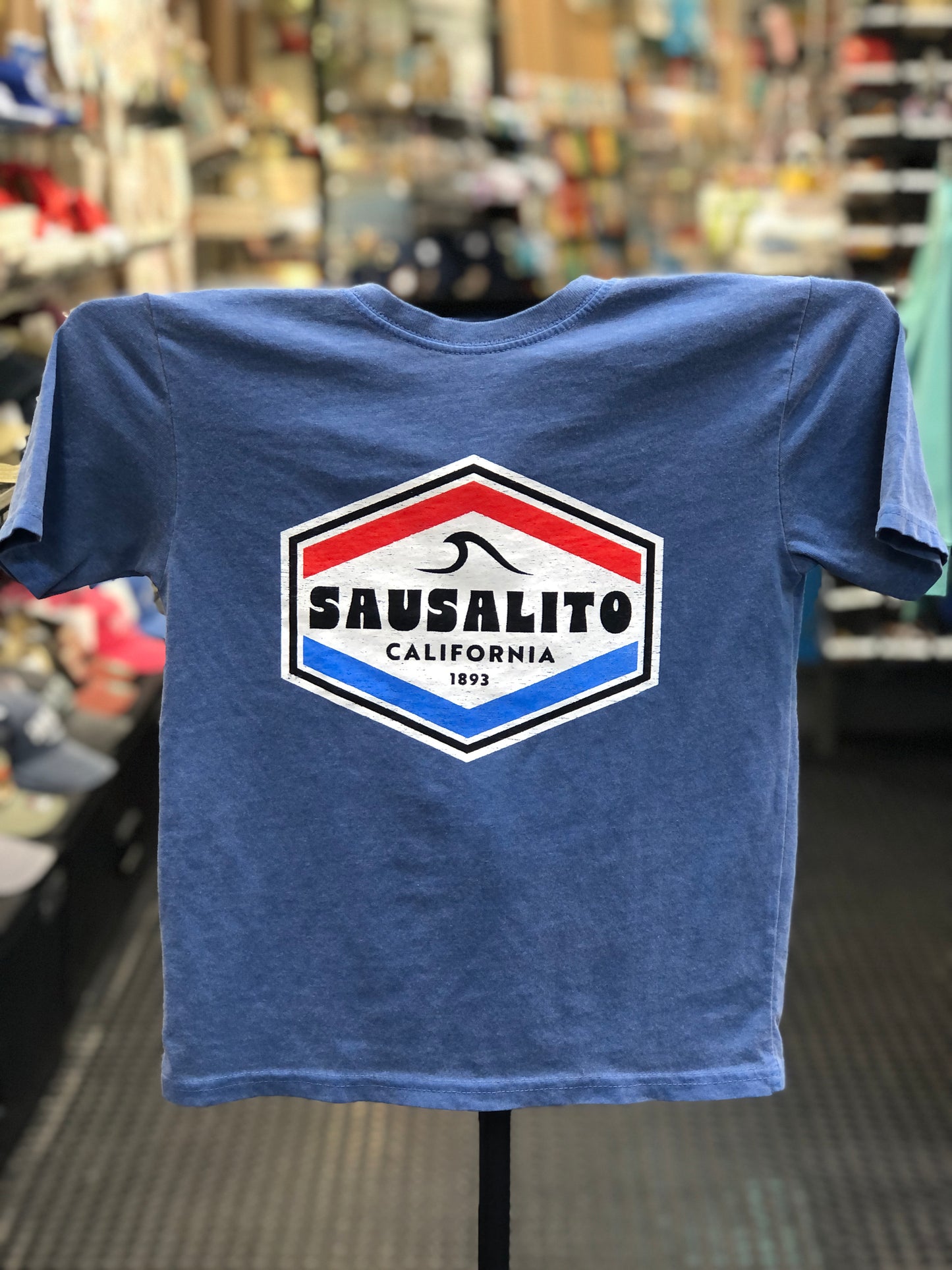 A blue heathered t-shirt featuring a retro red, white, and blue shield emblem with 'Sausalito California 1893' and a wave graphic, displayed on a clothing stand inside a shop.