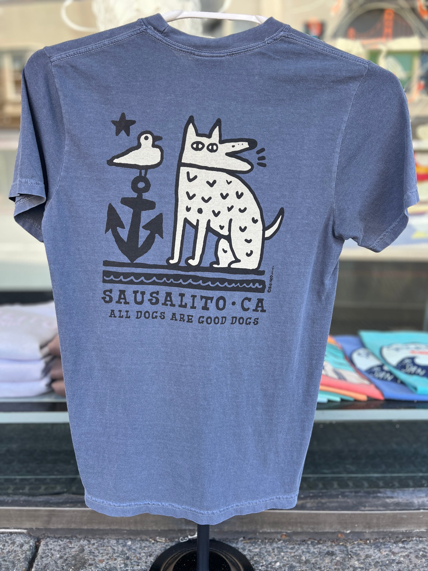 The back of a faded blue t-shirt displaying a white graphic of a stylized, spotted dog barking next to a large black anchor with a seagull perched on top. The words "Sausalito, CA" and "All Dogs Are Good Dogs" are printed beneath the design.