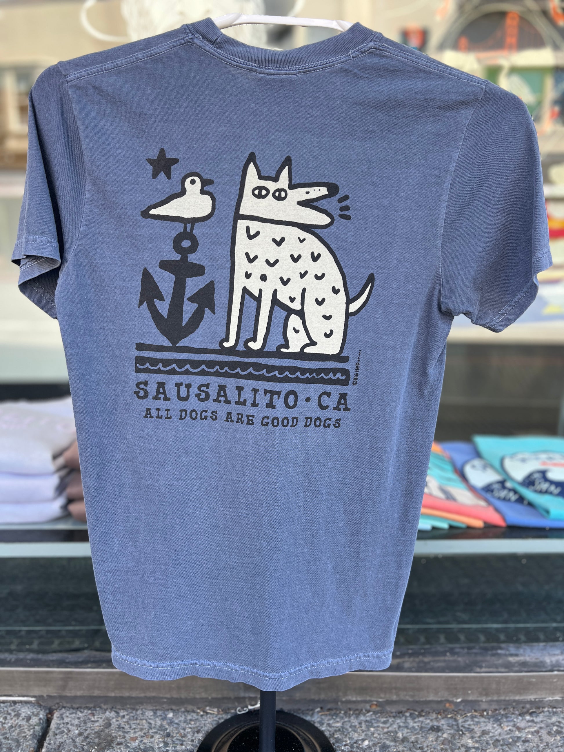 The back of a faded blue t-shirt displaying a white graphic of a stylized, spotted dog barking next to a large black anchor with a seagull perched on top. The words "Sausalito, CA" and "All Dogs Are Good Dogs" are printed beneath the design.