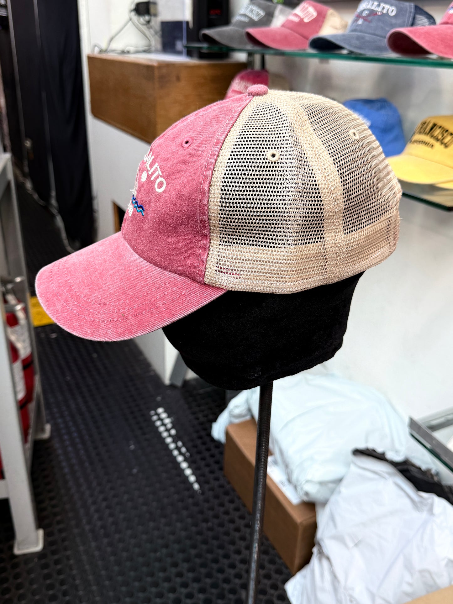 A side profile view of an unstructured trucker hat on a display stand inside a shop. The front panels and brim are a washed, pigment-dyed brick red cotton twill, while the back sections are a breathable tan mesh. Clean white embroidered lettering reading "SAUSALITO" arches across the front panel over a detailed pair of crossed rowing sculls.