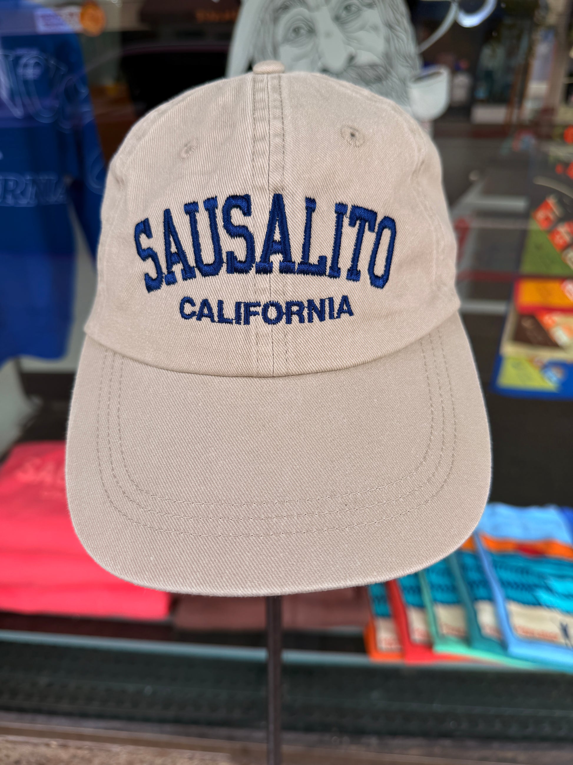 Front view of a khaki beige washed cotton baseball cap featuring "Sausalito" embroidered across the front in a bold navy blue arched font, with "California" stitched directly underneath.