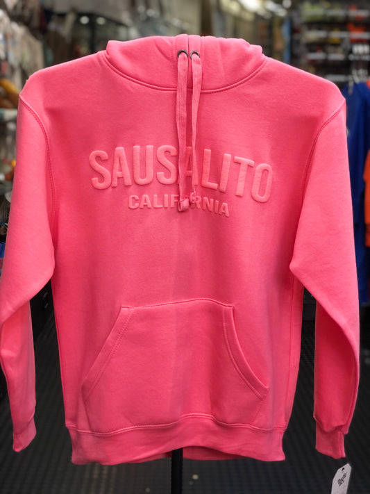 Bright rosy pink unisex hoody featuring a high-texture, raised puff-print graphic that reads "SAUSALITO CALIFORNIA" across the chest, styled with thick shoelace drawstrings and a clean crossover front pocket against a blurred background.