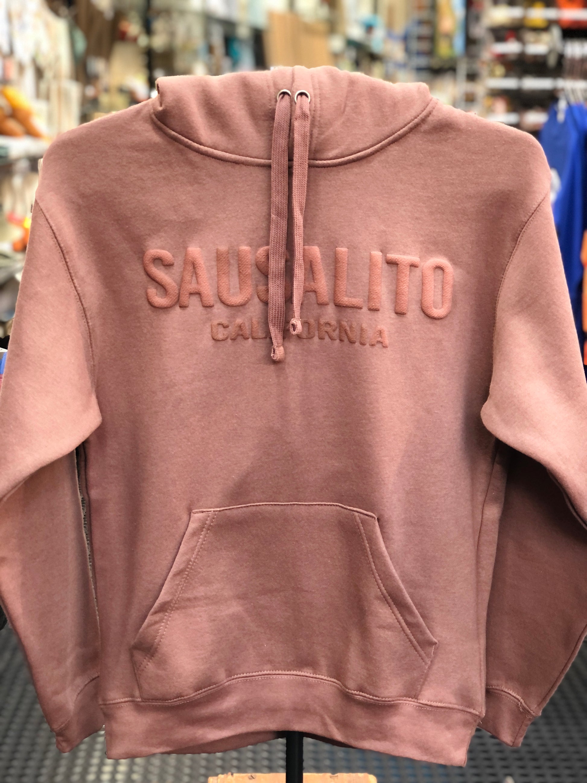 Front view of a clay color unisex hoody on a display stand inside a store, showcasing the prominent, raised puff-print lettering that reads "SAUSALITO CALIFORNIA" across the chest with matching thick hood drawstrings.