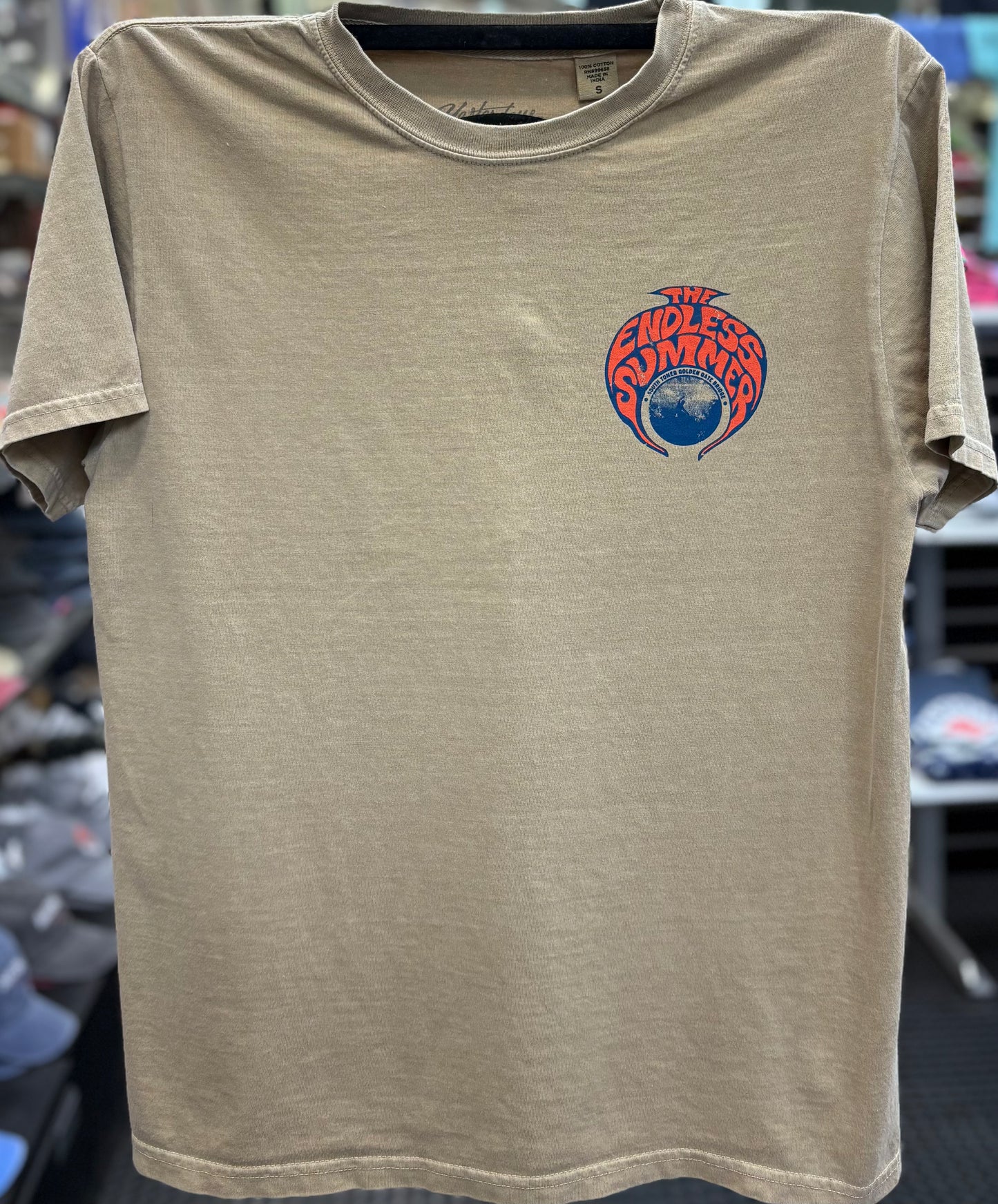 Front view of a khaki-colored garment-dyed crewneck t-shirt hanging on a clothing rack. The shirt features a vibrant circular graphic screen-printed on the left chest with an orange and blue psychedelic frame surrounding a central illustration of a surfer riding a wave beneath the South Tower of the Golden Gate Bridge, accented by the text "THE ENDLESS SUMMER".