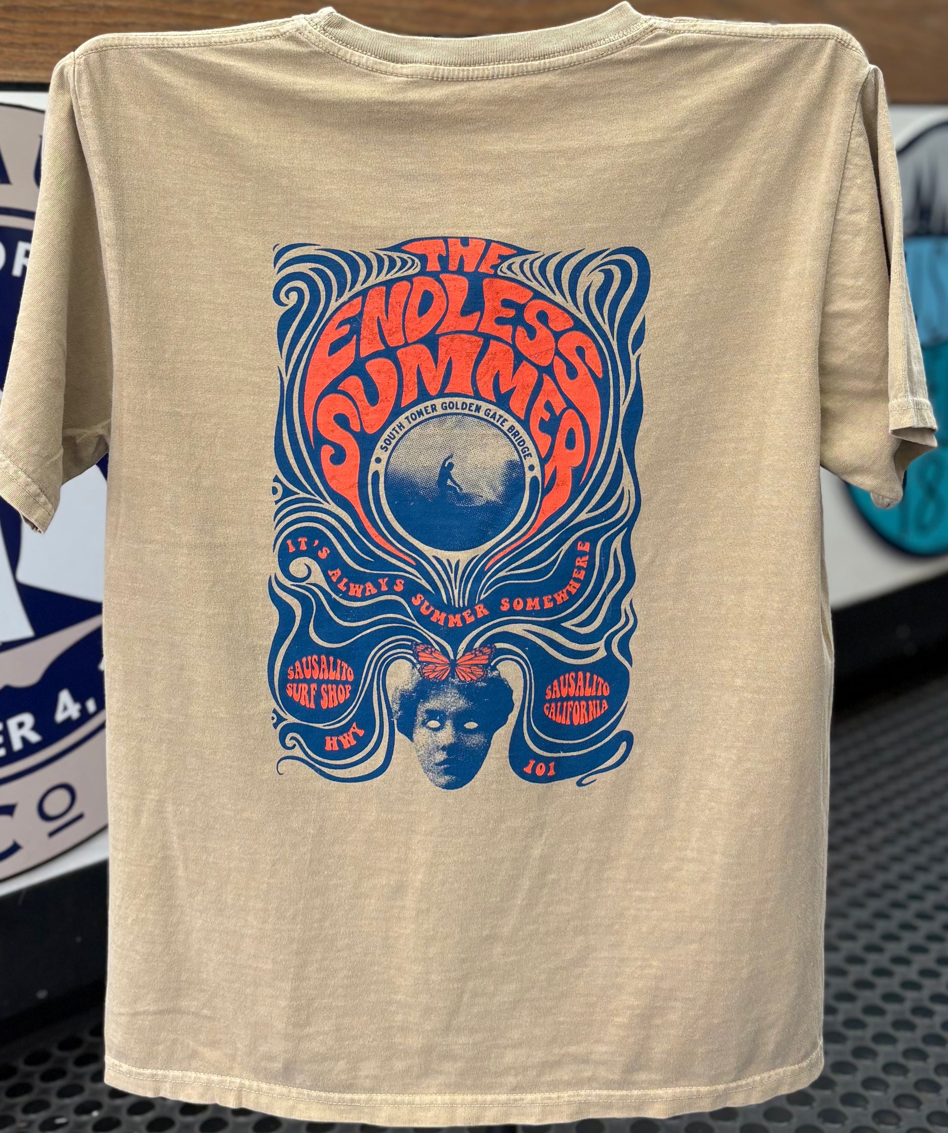 Back view of a khaki-colored garment-dyed crewneck t-shirt. The shirt features a large, detailed screen-printed graphic in vibrant orange and blue showcasing psychedelic, flowing wavy lines that frame a central circular image of a surfer riding a wave beneath the South Tower of the Golden Gate Bridge. The stylized text reads "THE ENDLESS SUMMER" across the top, with "IT'S ALWAYS SUMMER SOMEWHERE" along the circle, and "SAUSALITO SURF SHOP • HWY 101 • SAUSALITO CALIFORNIA" anchoring the bottom design around 
