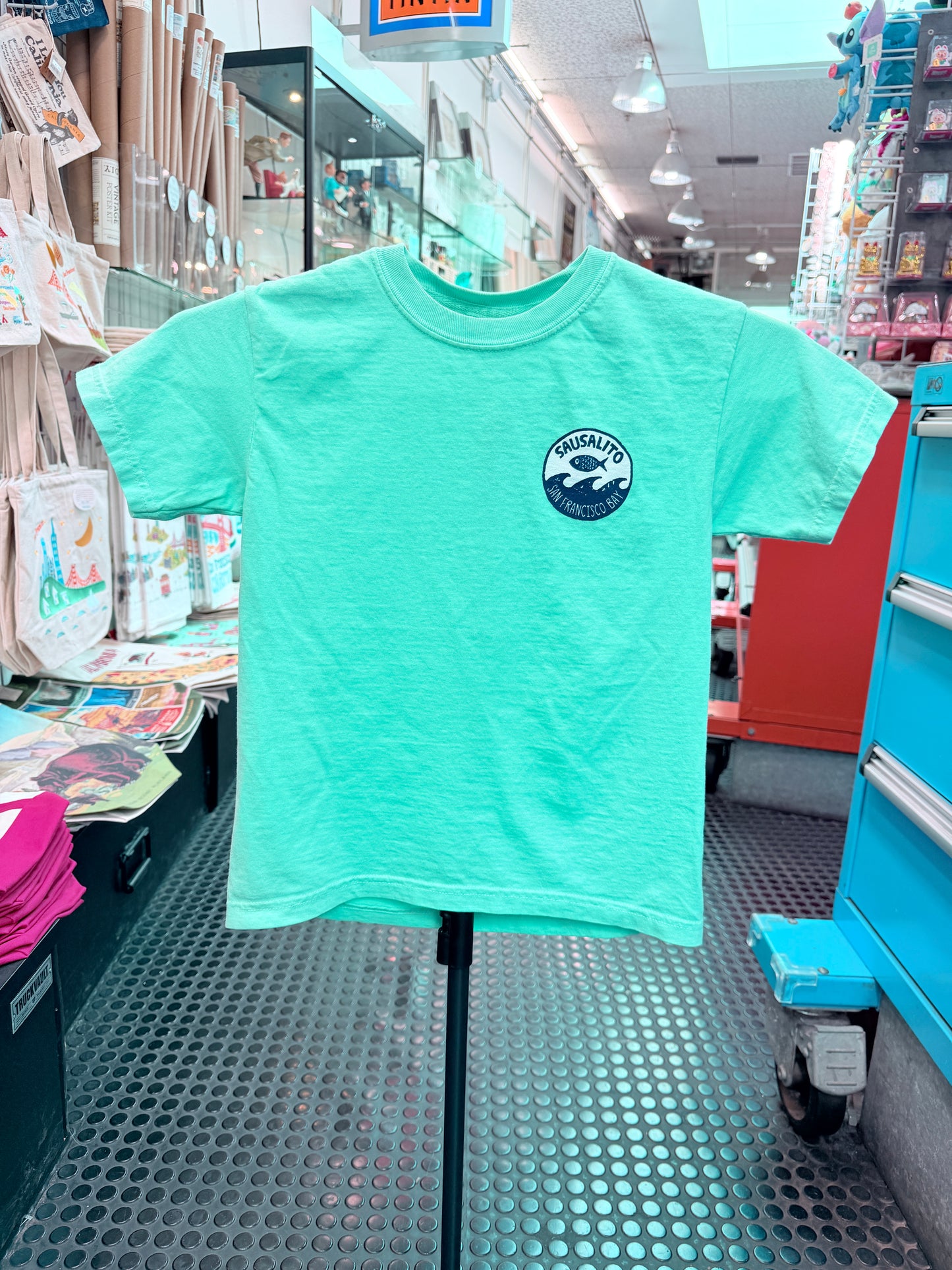 Front of a mint green short-sleeve kids T-shirt displayed on a stand inside the Sausalito Ferry Co. retail store. The left chest features a small circular navy blue and white logo showcasing a fish jumping over ocean waves, surrounded by the text "Sausalito" and "San Francisco Bay."